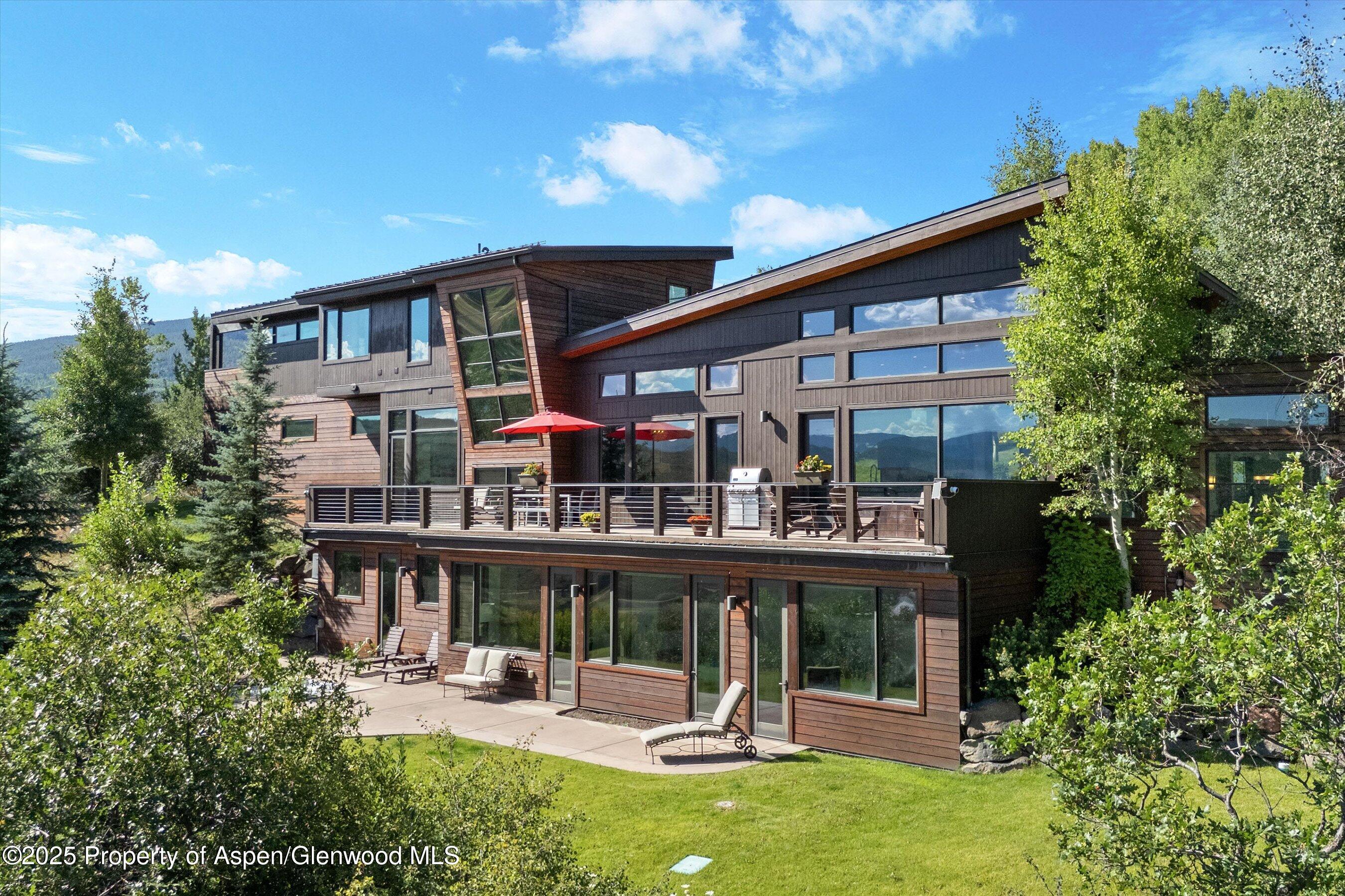 Modern wood-sided home with a large deck and mountain views, surrounded by trees and lawn.