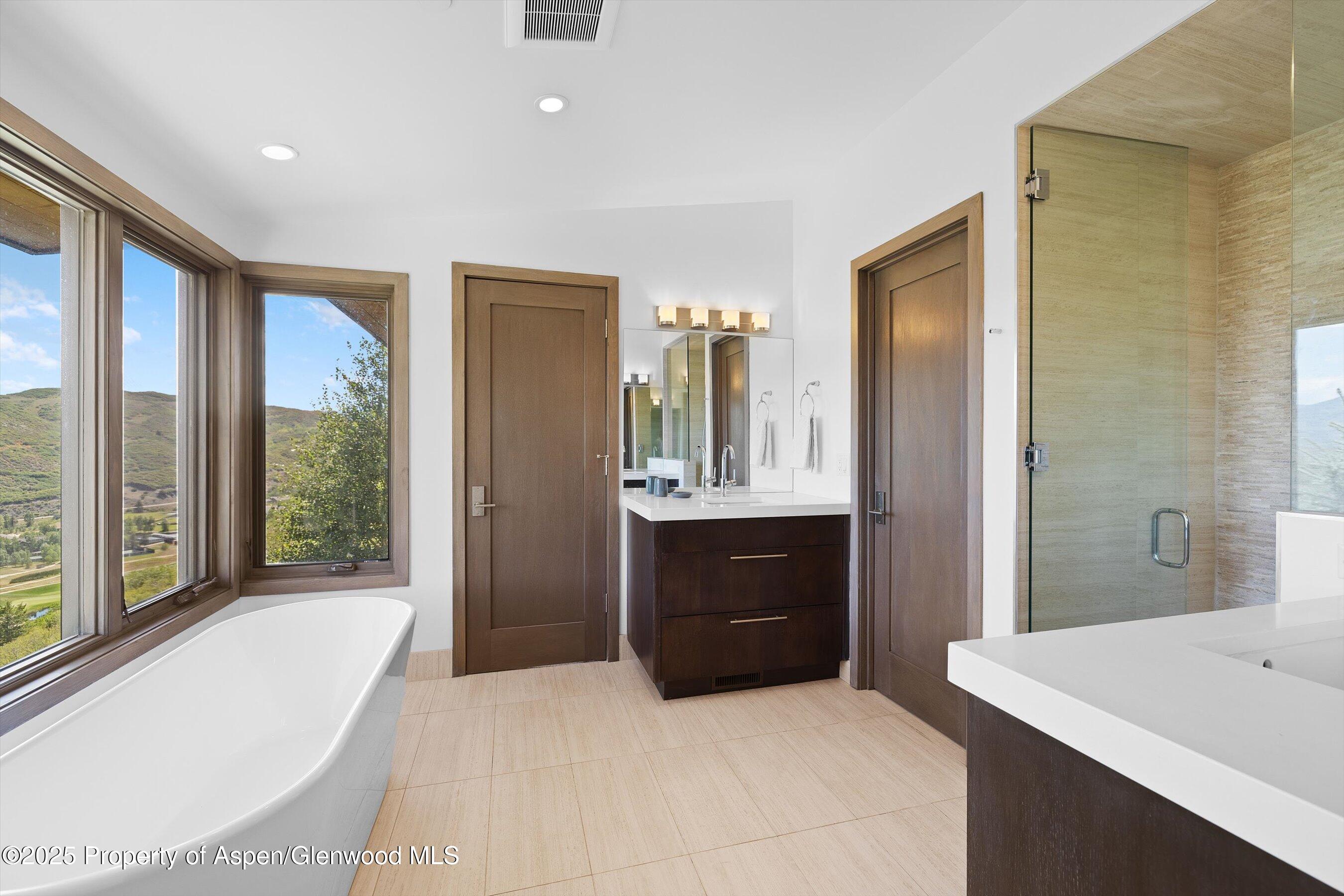 Modern bathroom with a freestanding tub, glass shower, dark wood vanity, and hillside views through large windows.