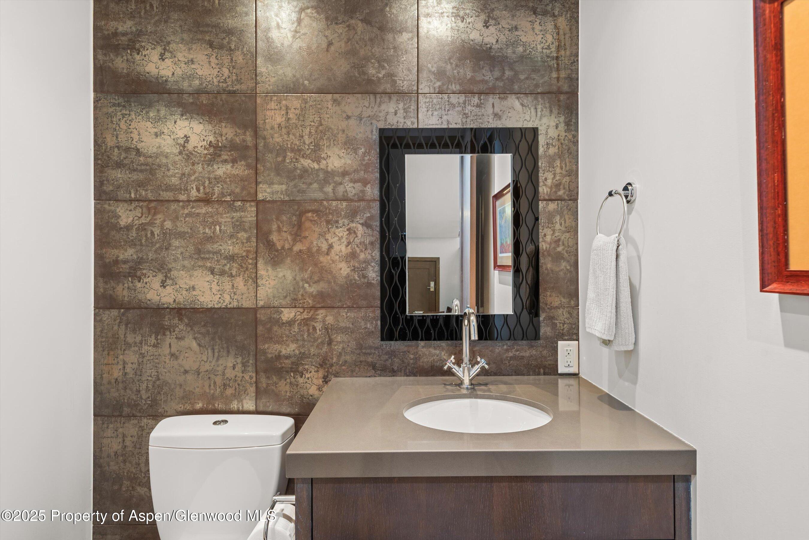 Bathroom vanity and toilet against a textured brown tile wall, with a black-framed mirror.