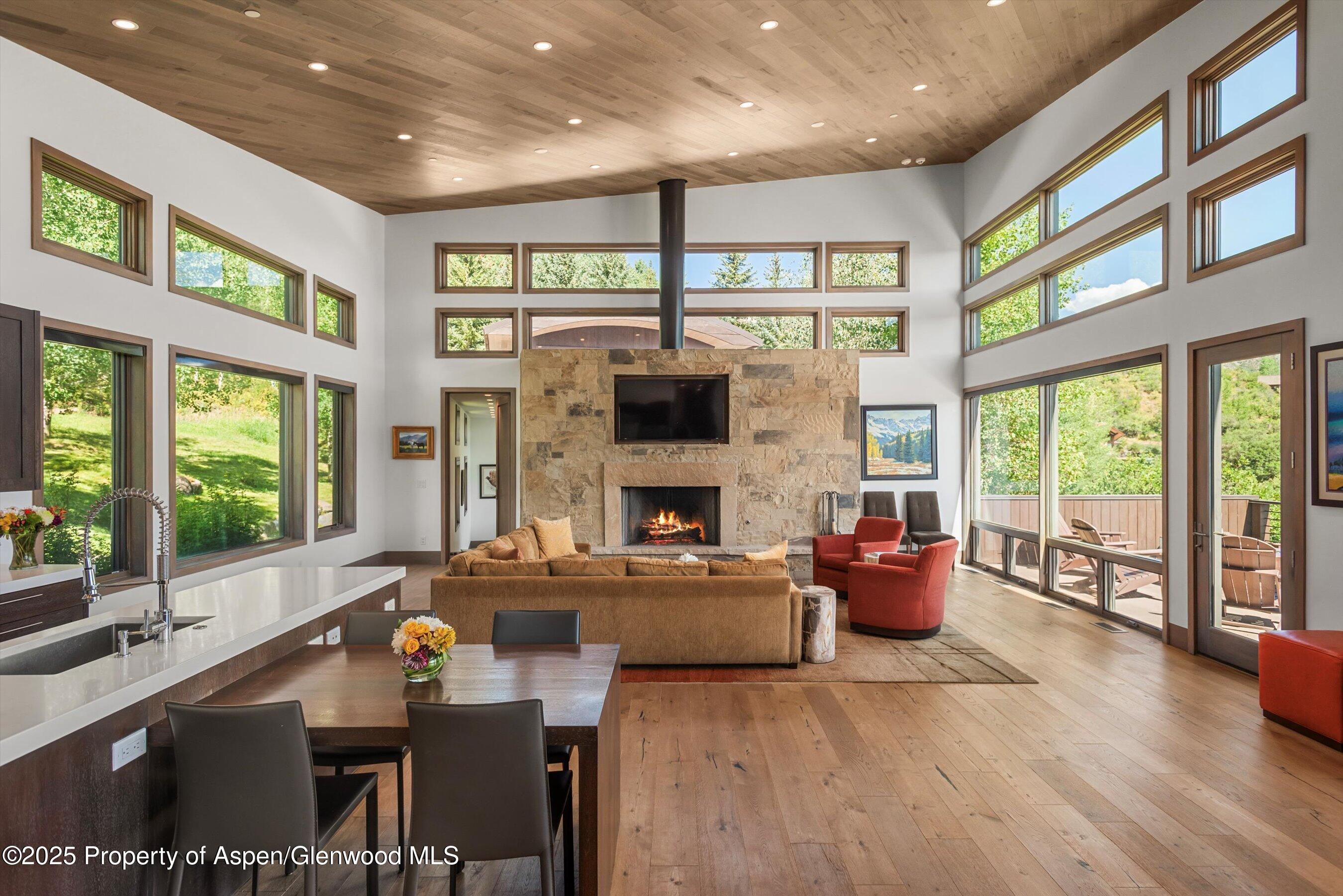 Open-plan living room with a stone fireplace, soaring wood ceiling, and tall windows overlooking greenery.