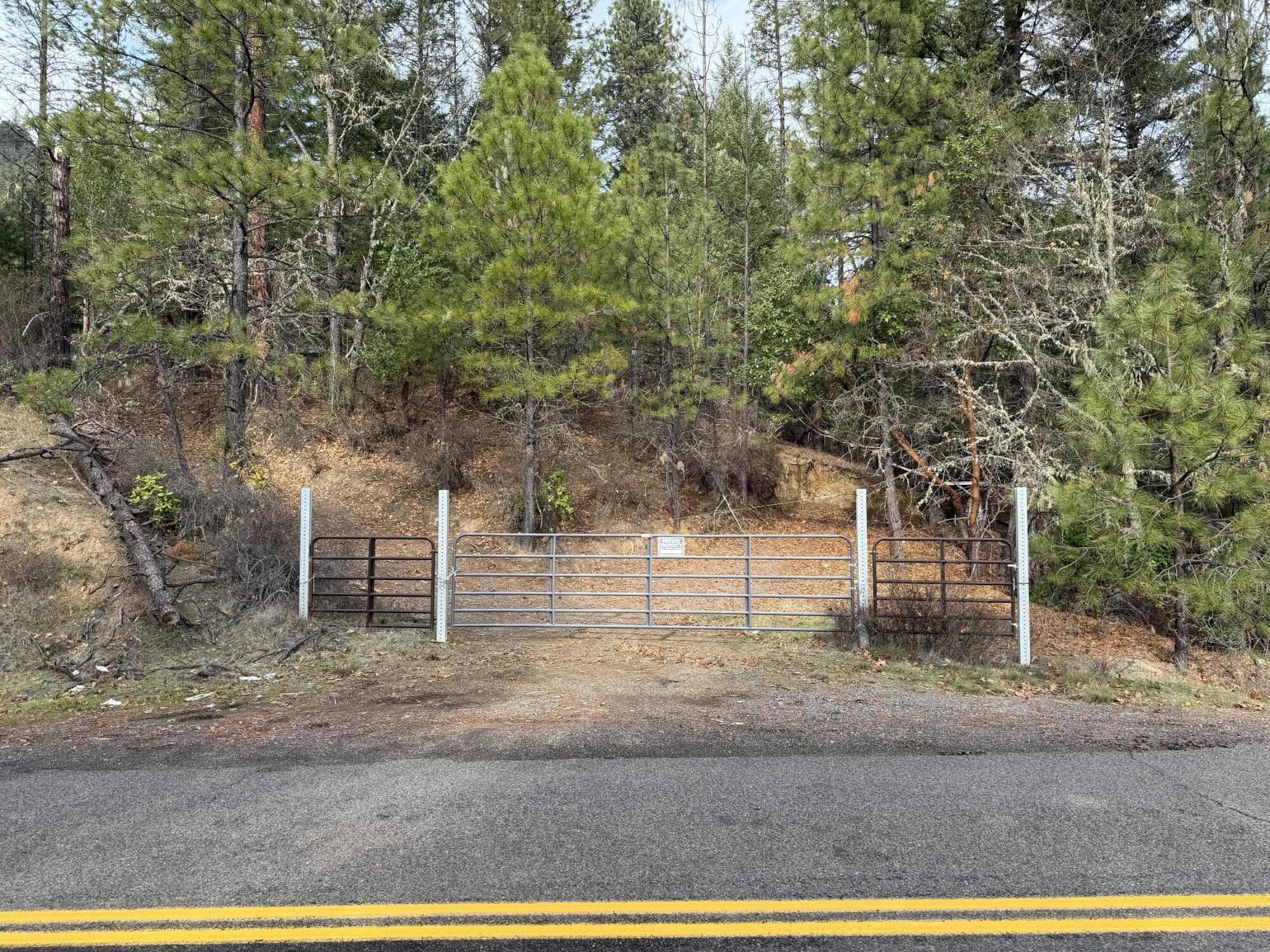 Wooded vacant lot behind a metal gate, with a dirt driveway opening onto a paved road.