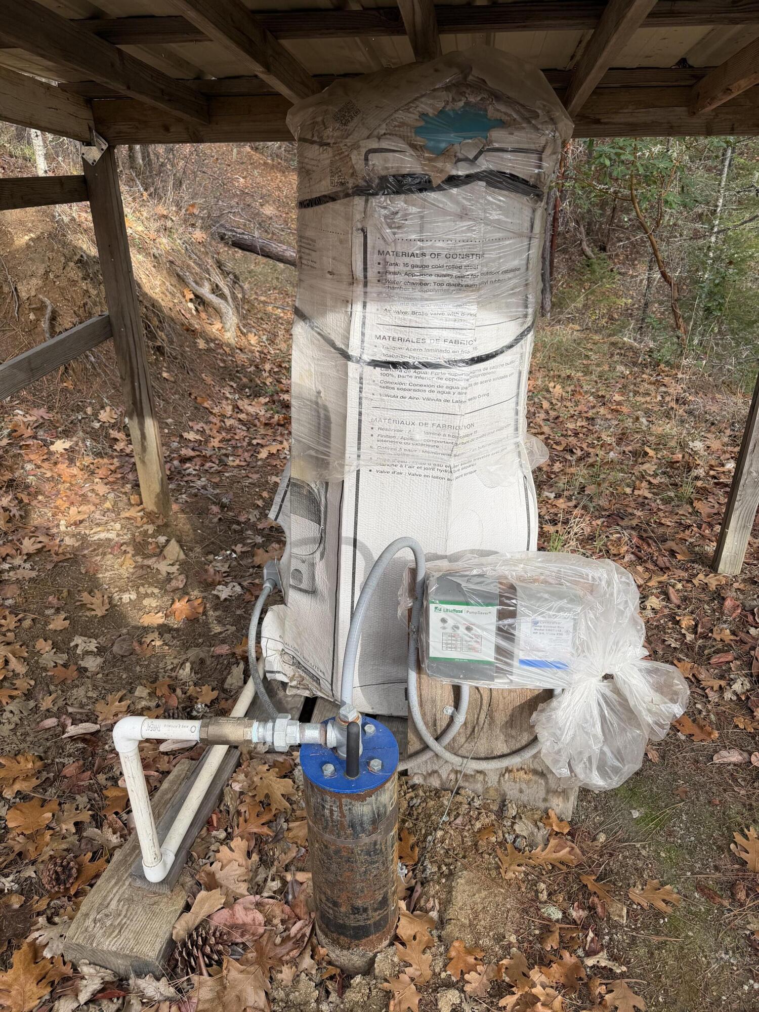 Well pump and wrapped pressure tank beneath a wooden shelter, with pipes connected to the well casing.
