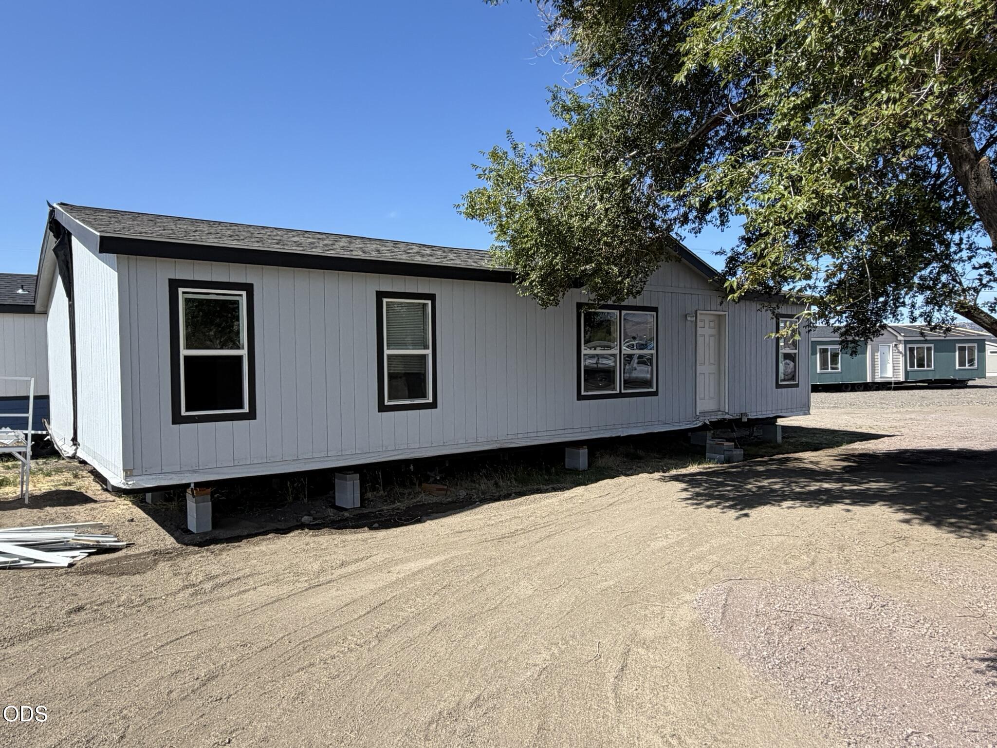 Light gray manufactured home raised on support blocks beside a dirt driveway.