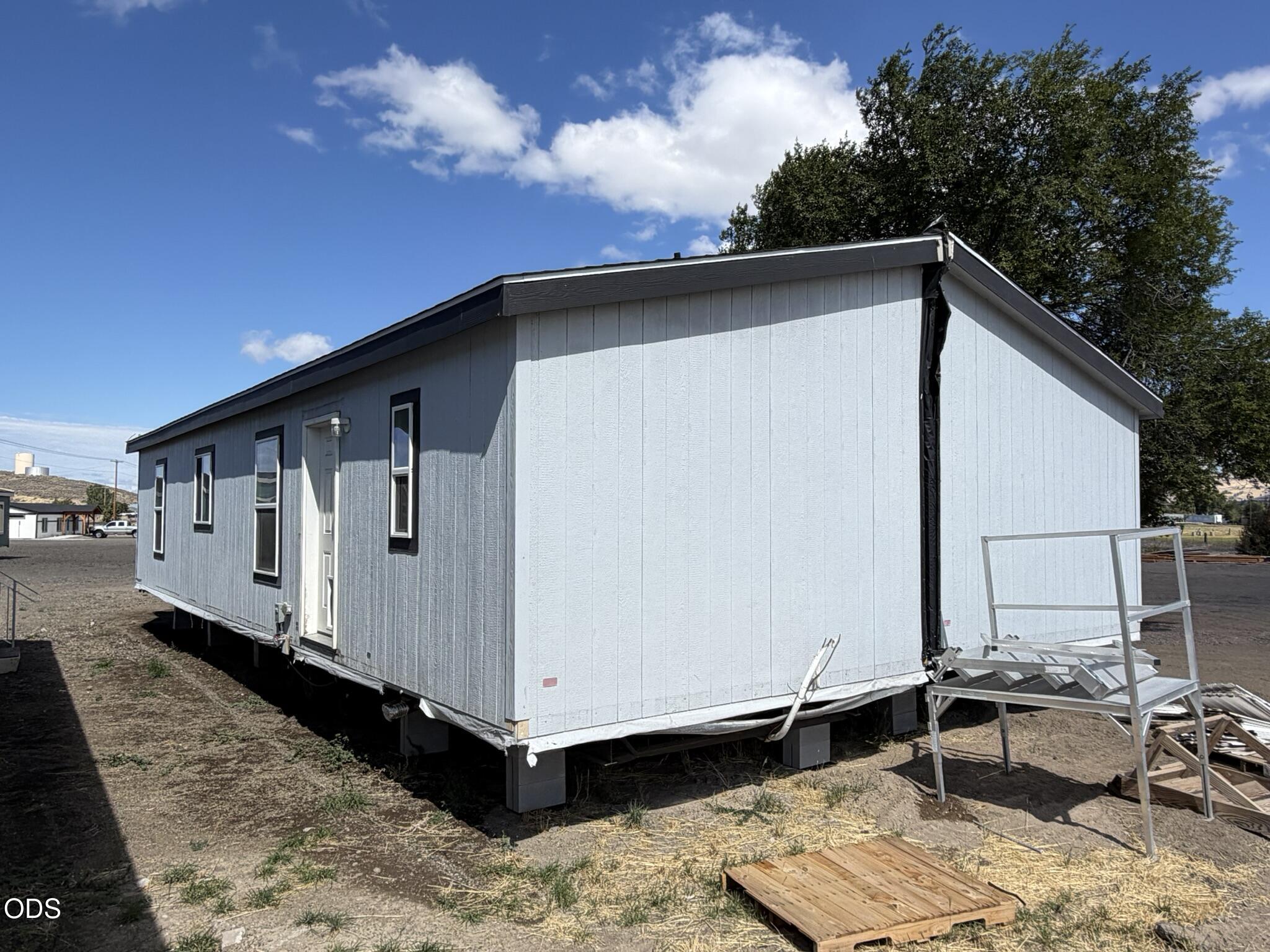 Light-colored manufactured home raised on supports, with loose stair framing and a wooden platform beside it.