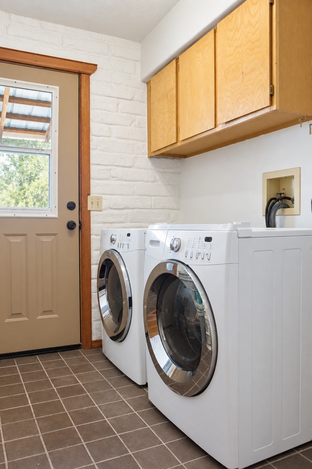 Laundry room with front-loading washer and dryer beneath light wood cabinets beside a windowed door.