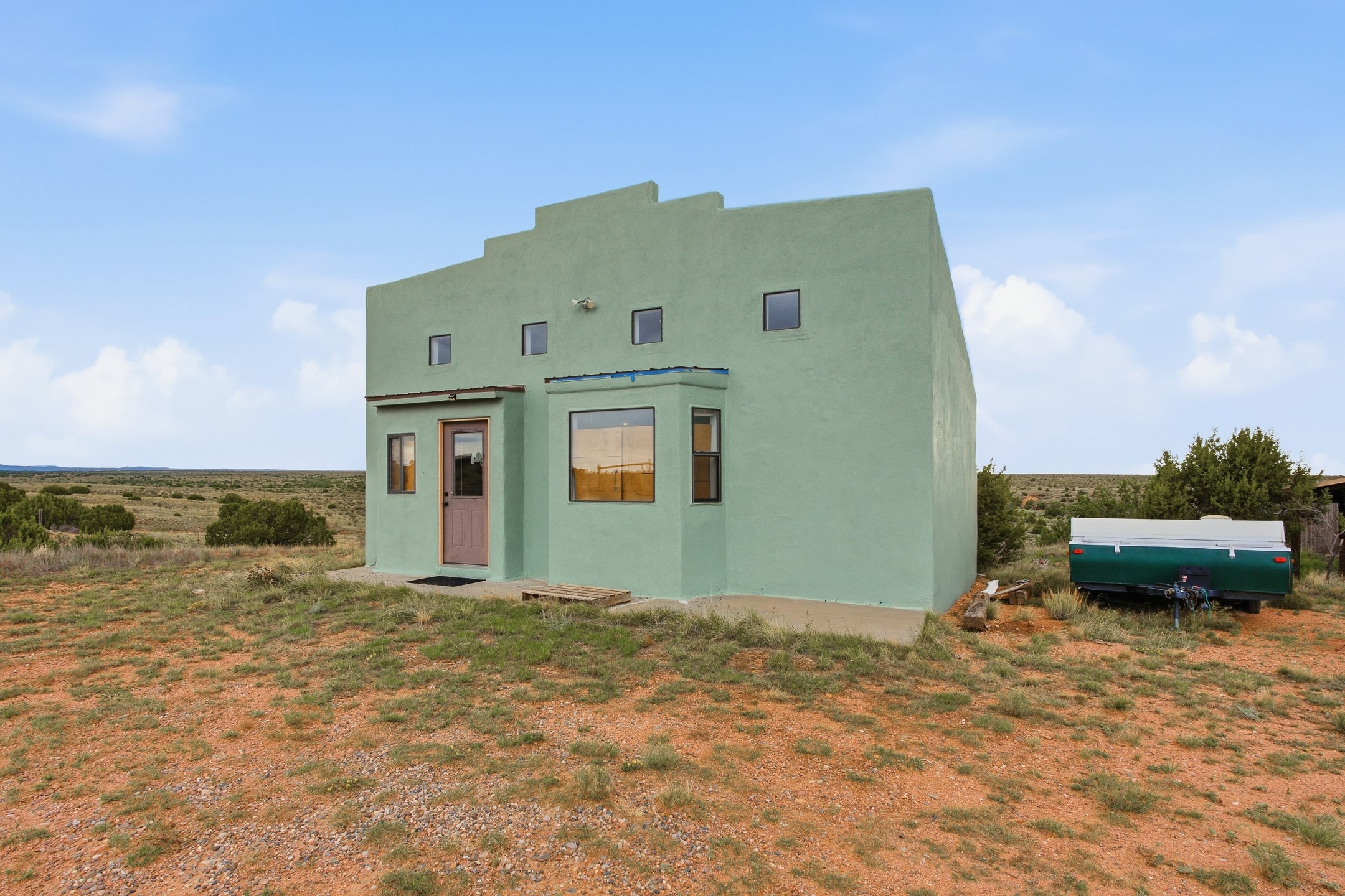 Sage-green stucco home with stepped parapets and a pink door on open scrubland beside a trailer.