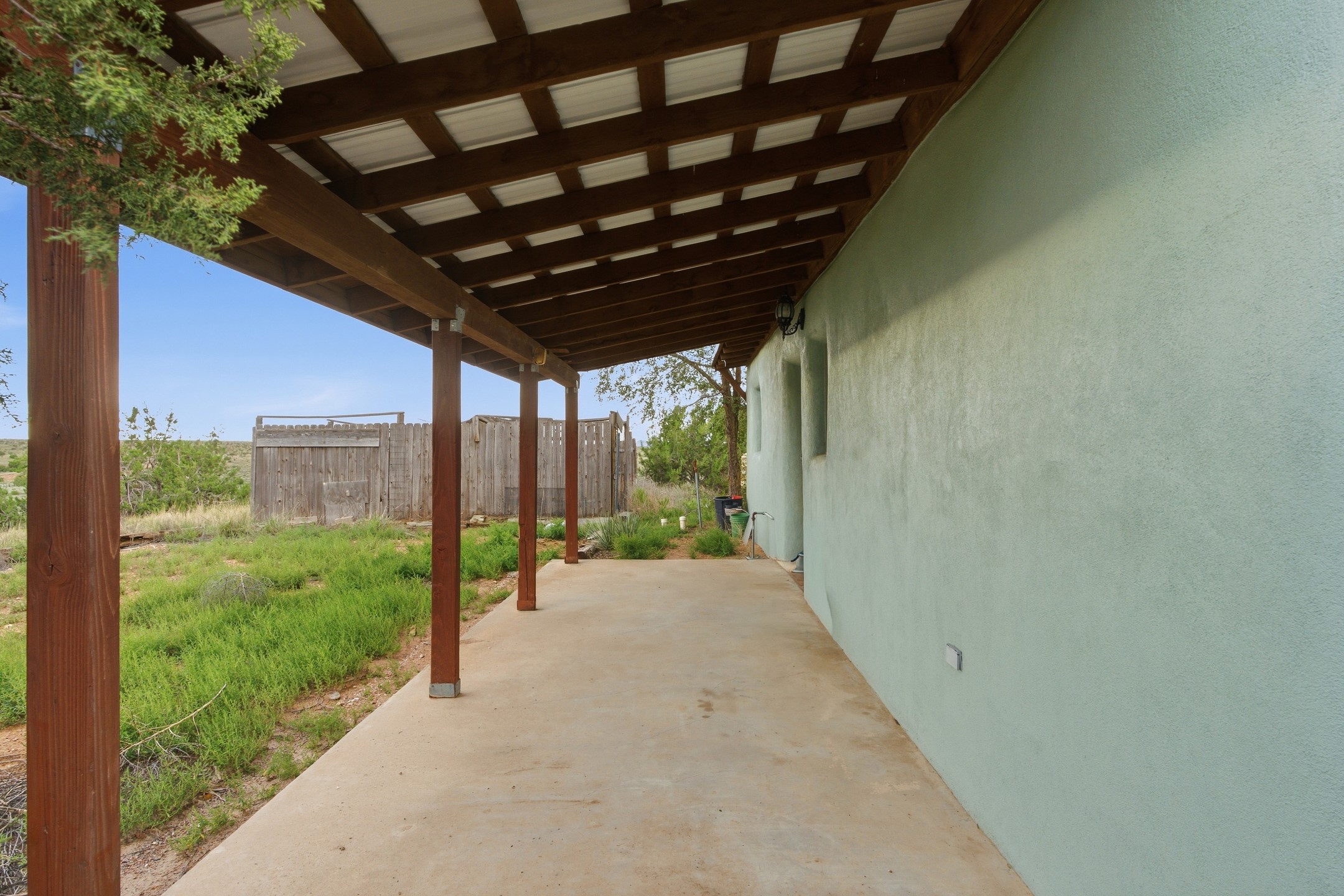 Covered concrete patio beside a stucco house, with timber posts and a fenced yard.