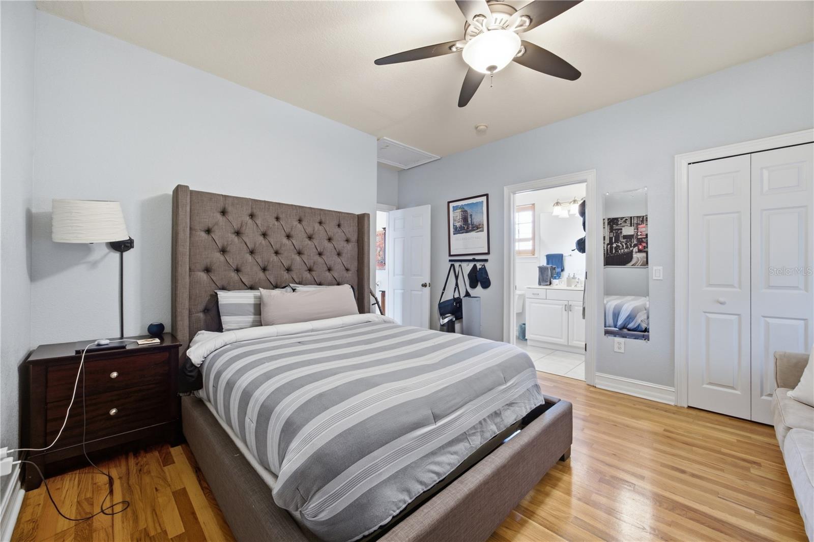 Bedroom with a tall tufted headboard, striped bedding, hardwood floors, and an adjoining bathroom.