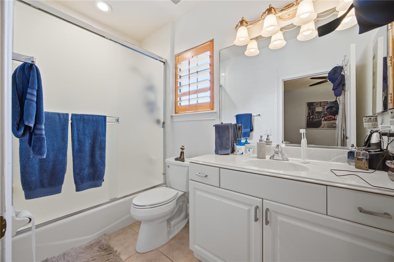 Bathroom with a glass-enclosed tub, white vanity, and blue towels beneath a shuttered window.