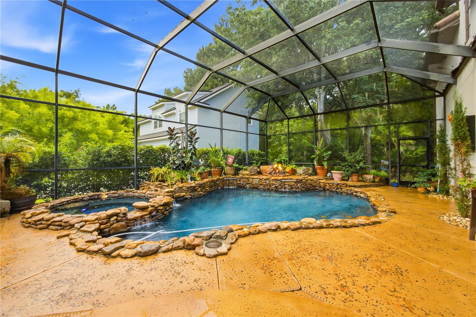 Screened backyard pool with a natural stone edge, rock waterfall, and potted tropical plants.