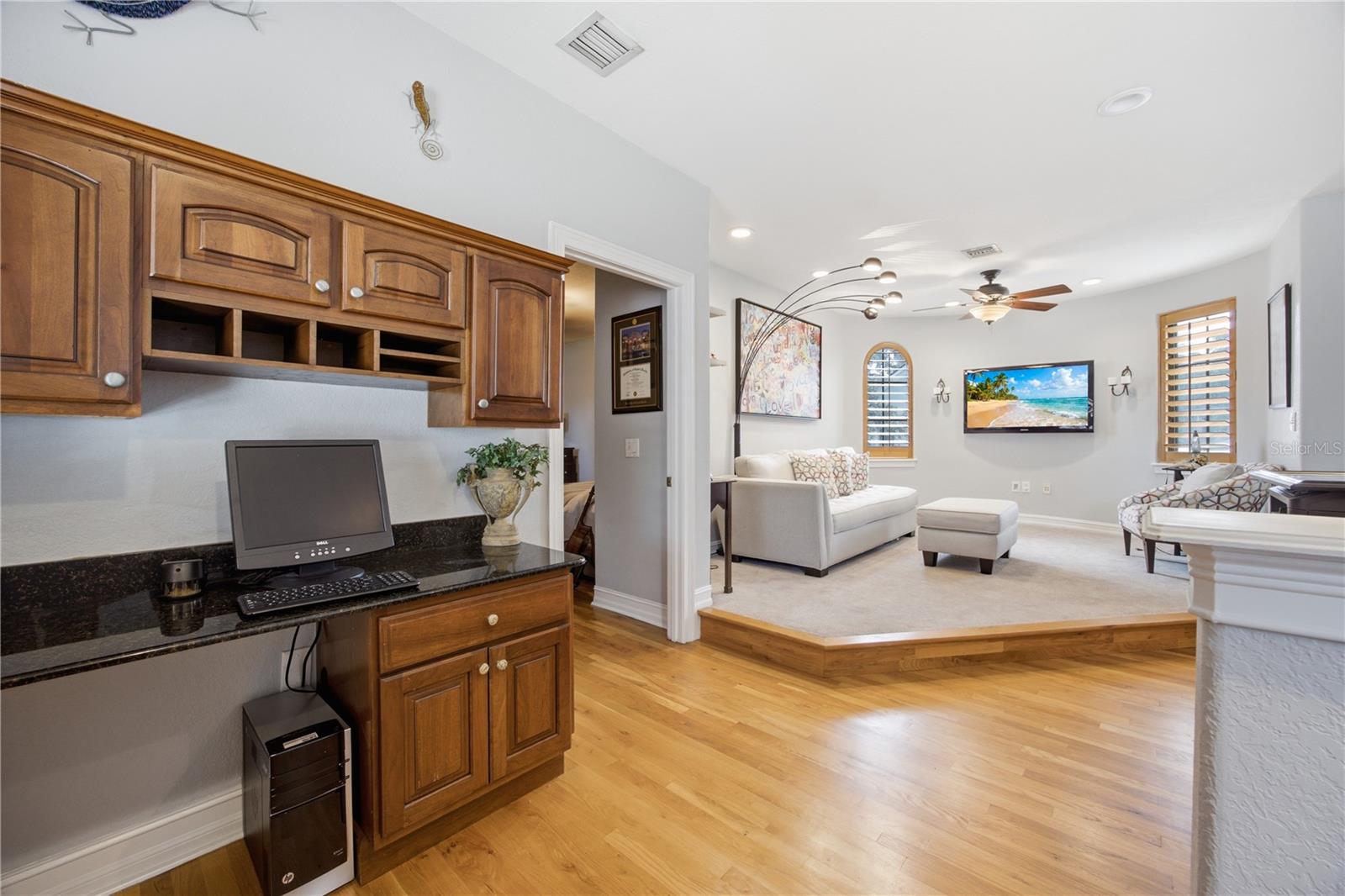 Home office with built-in wood cabinetry opens to a raised living room with white seating and plantation shutters.