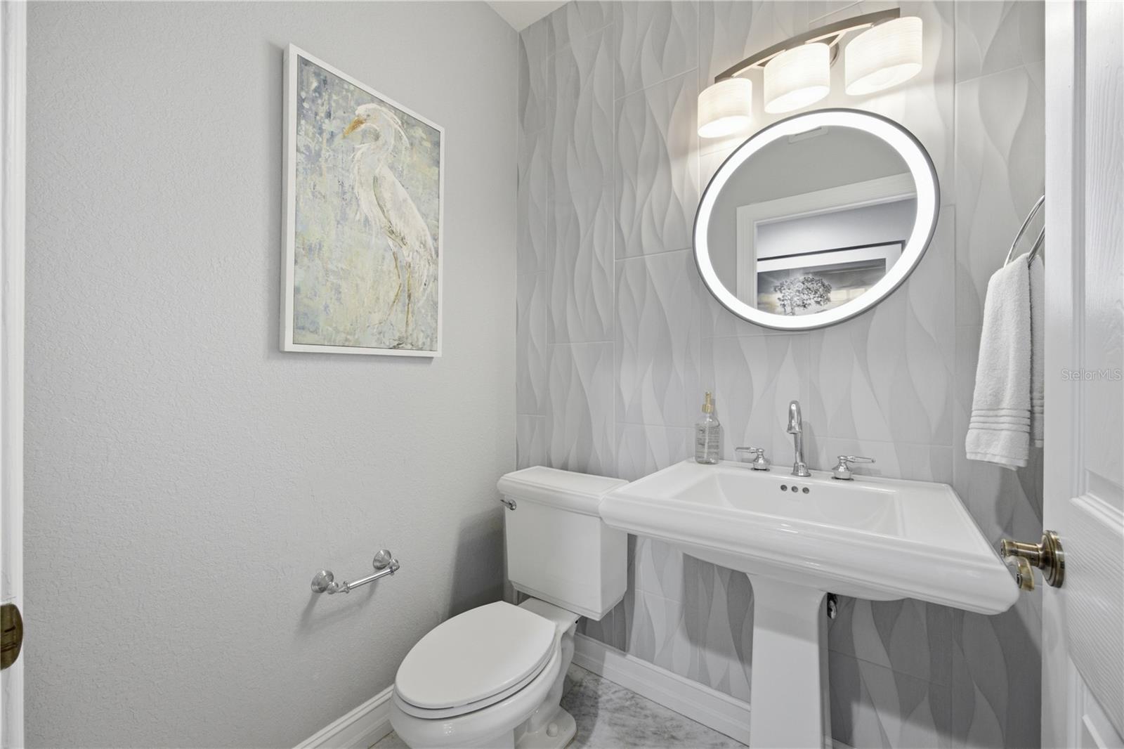 Bathroom with a white pedestal sink, illuminated oval mirror, and sculpted gray tile wall.
