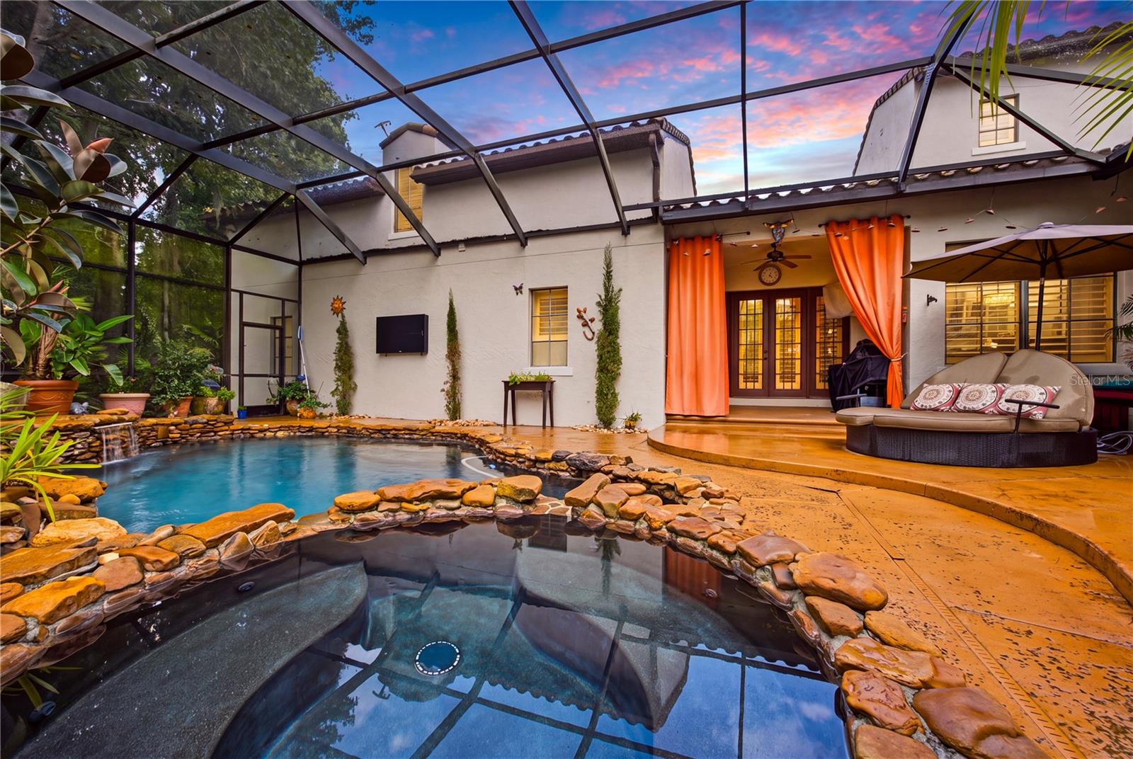 Backyard pool and raised spa beside a screened patio with orange curtains at sunset.