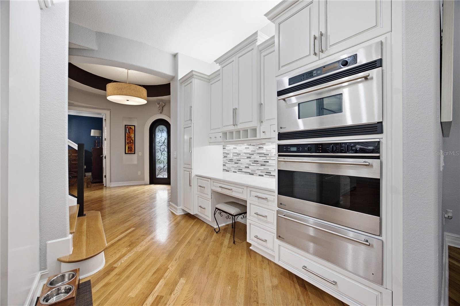 Kitchen with white cabinetry, stacked stainless-steel ovens, mosaic backsplash, and hardwood floors.