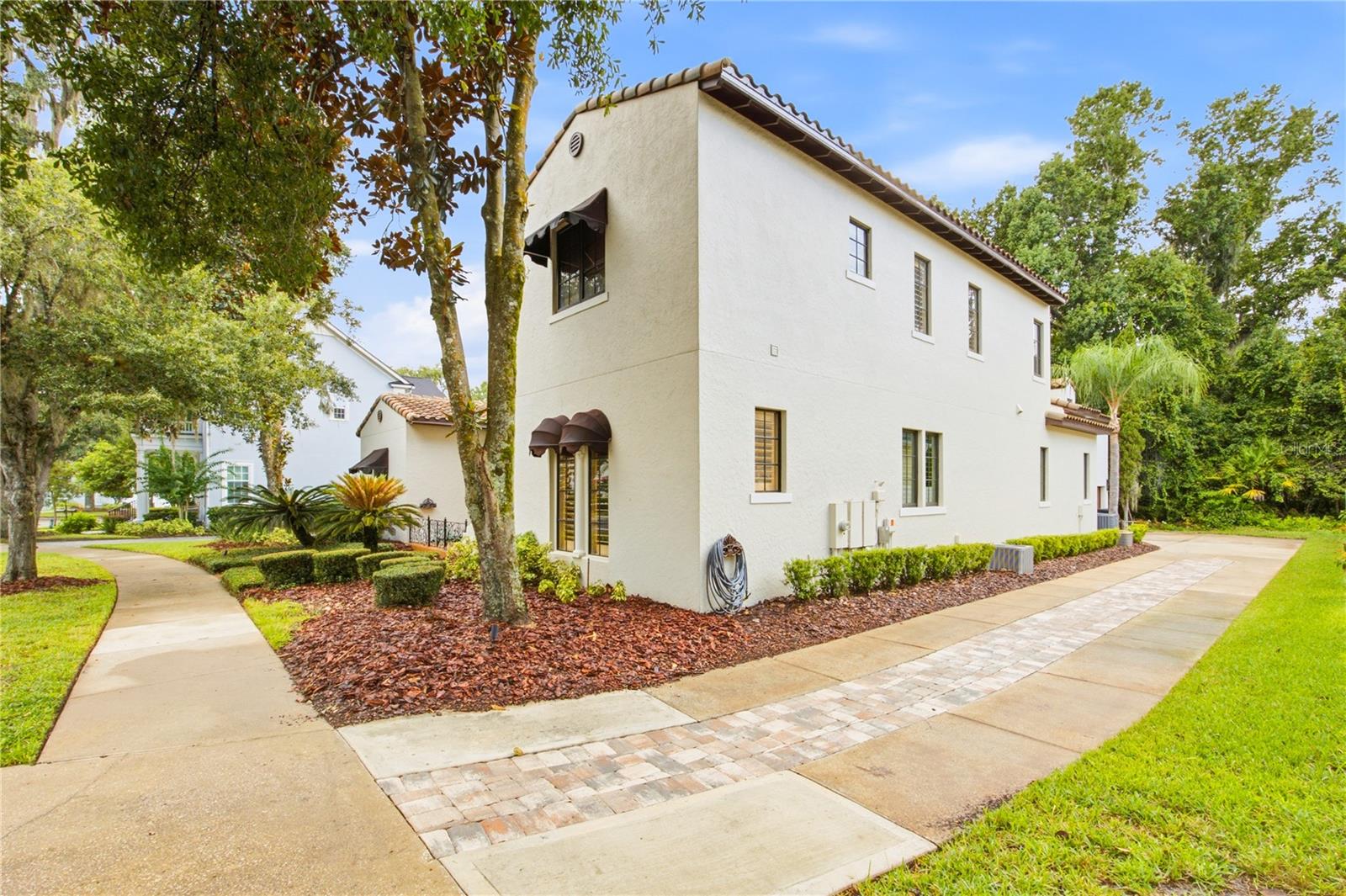 Two-story cream stucco home with a tile roof, dark awnings, and landscaped grounds.
