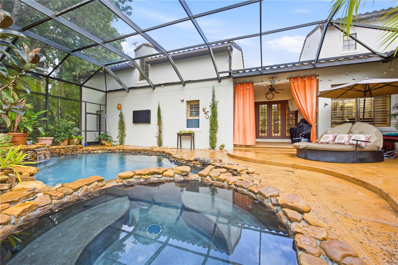 Backyard pool and spa with a stone border, screened enclosure, and raised patio with orange curtains.