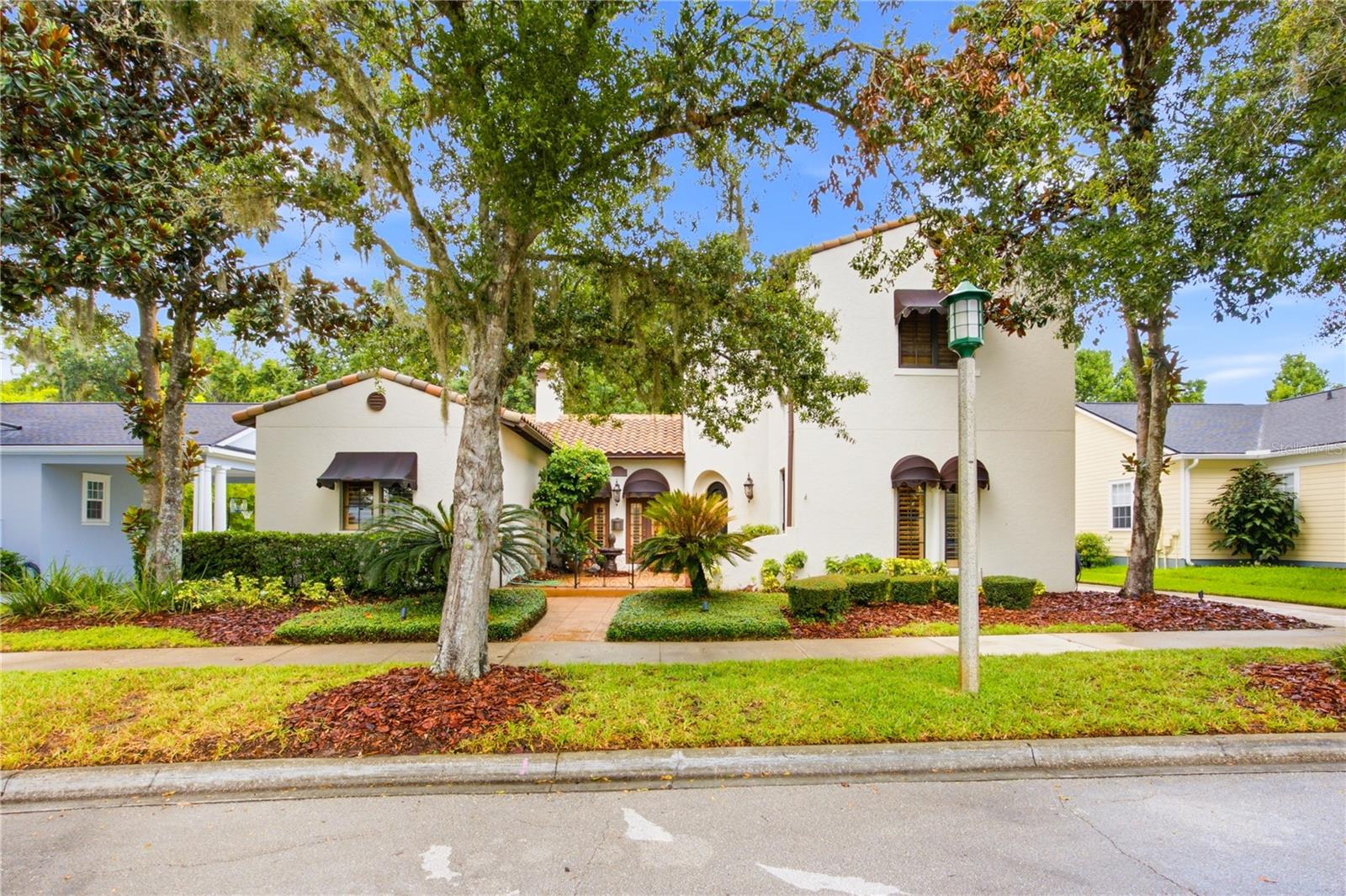 Front exterior of a cream stucco home with terracotta tile roofs and mature shade trees.