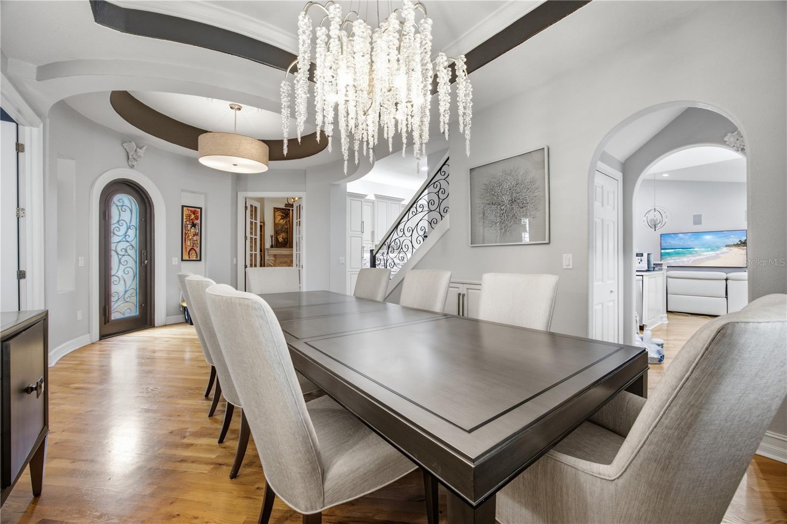 Dining room with a dark wood table, crystal chandelier, parquet flooring, and ornate staircase railing.