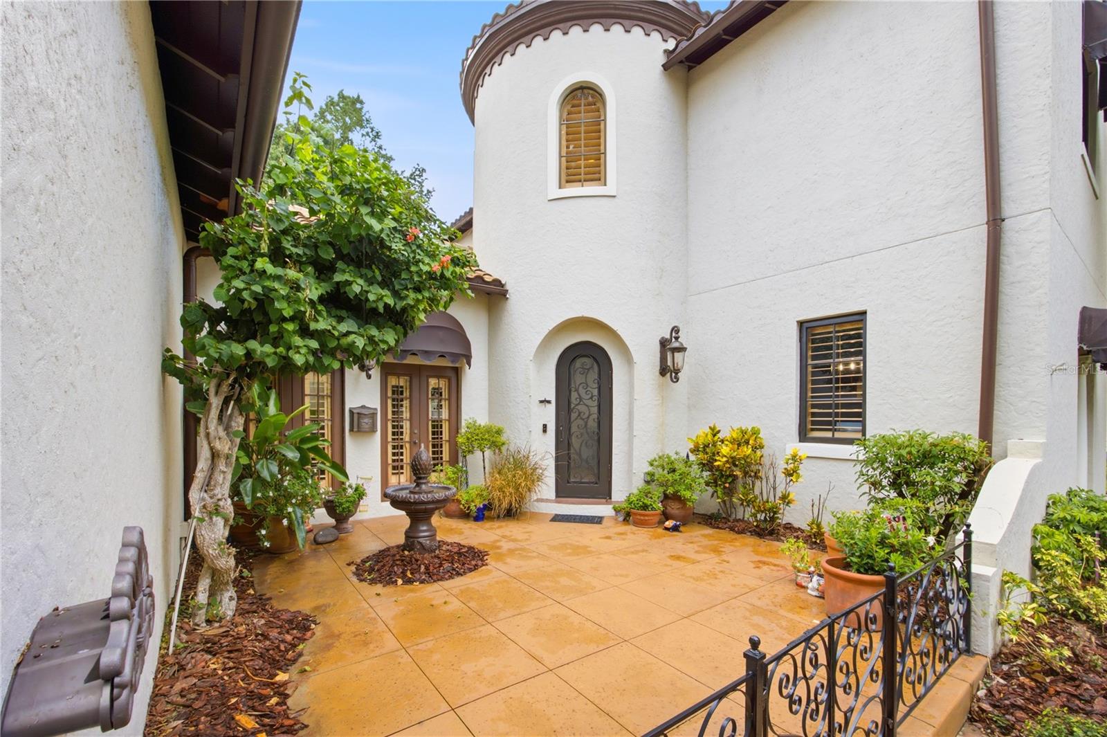 White stucco home entrance with an arched front door, tiled courtyard, fountain, and potted plants.