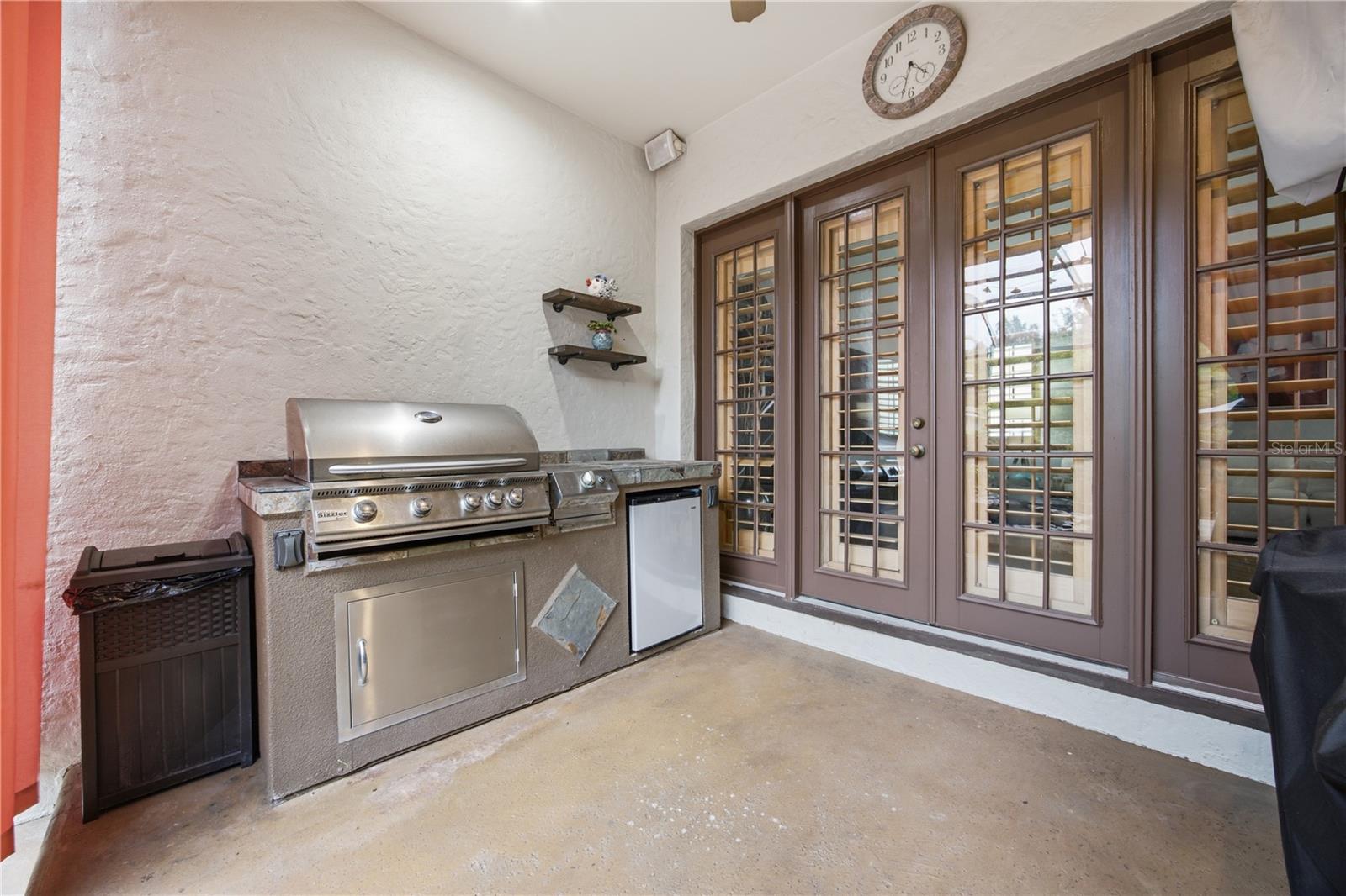 Covered patio with a stainless-steel grill and refrigerator beside brown-framed glass doors.