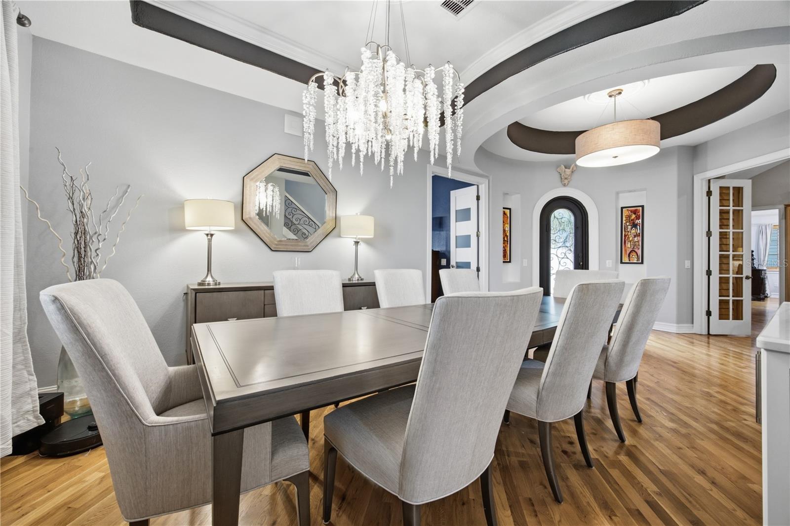 Dining room with a crystal chandelier, upholstered chairs, hardwood floors, and an arched front door.