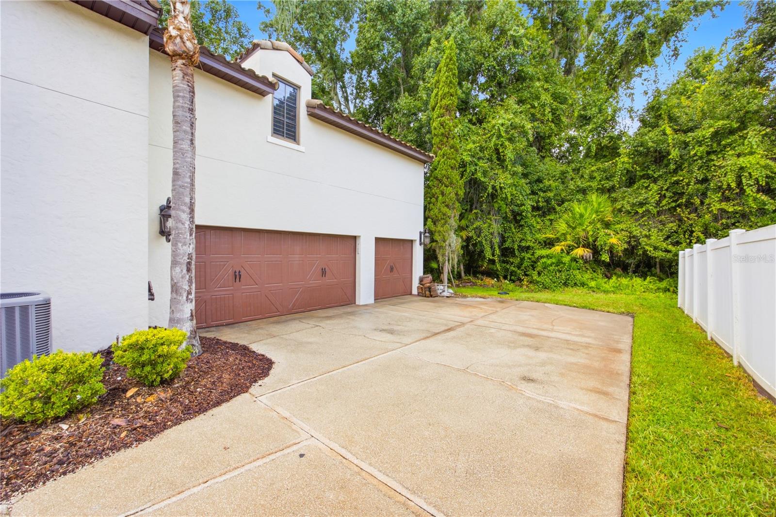 Home exterior with a wide concrete driveway, two brown garage doors, and a white fence.