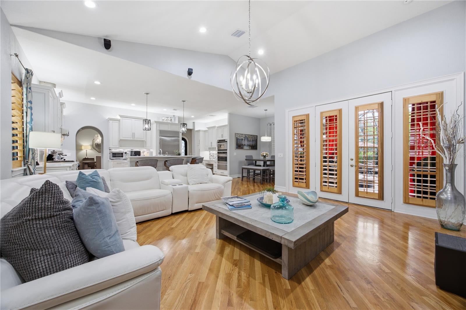 Open-plan living room with white sectional, hardwood floors, adjoining kitchen, and shuttered French doors.
