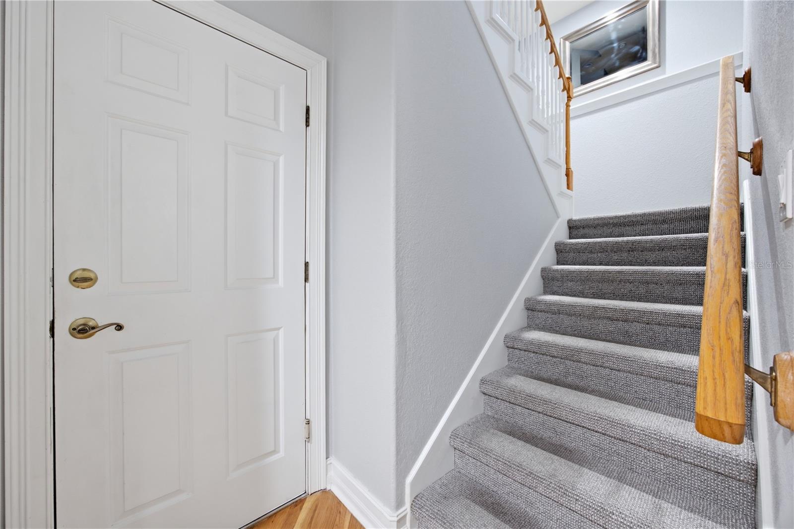 Entryway with a white paneled door beside a gray-carpeted staircase and wooden handrail.