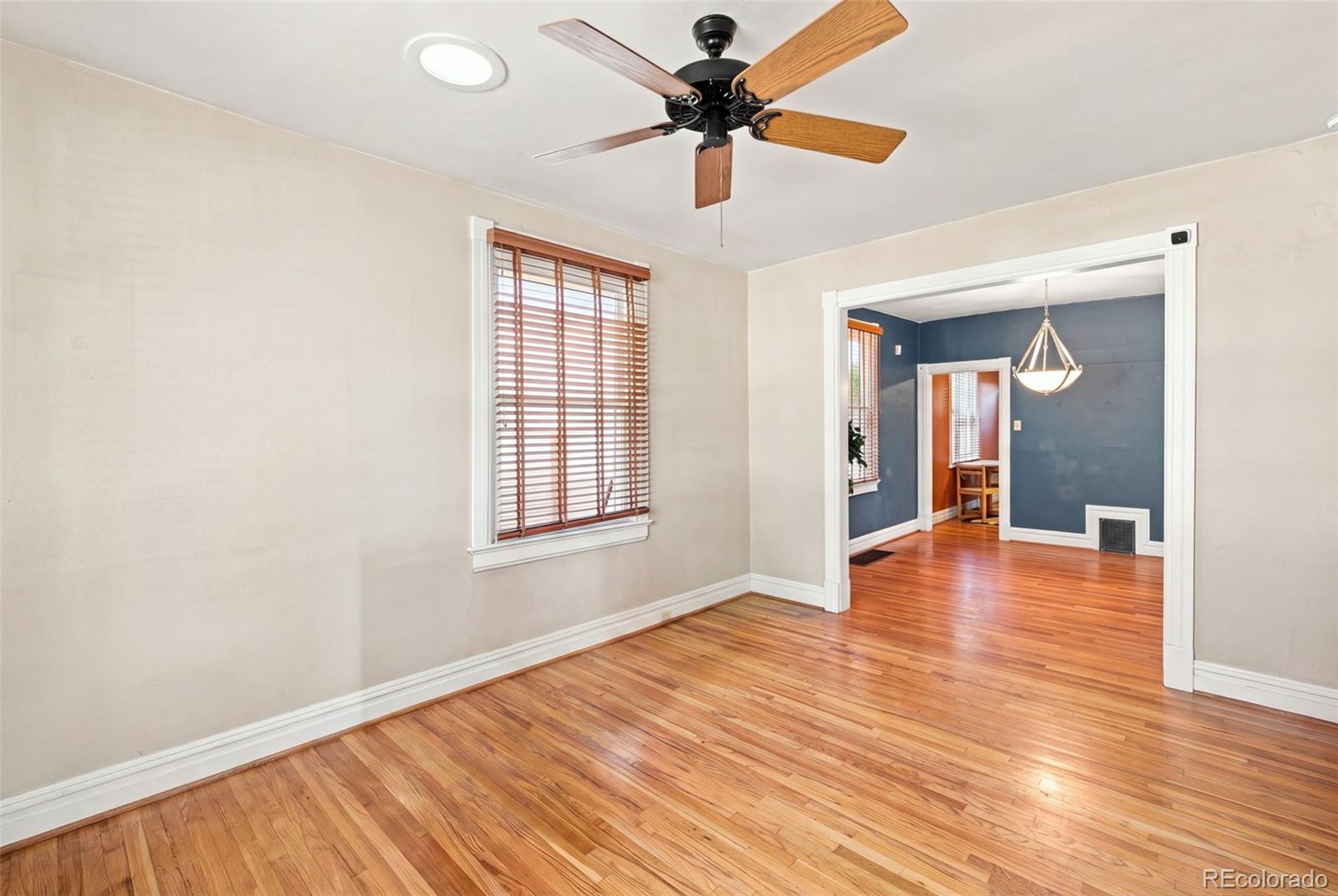 Empty living room with hardwood floors, a ceiling fan, and an opening to a blue-walled dining room.