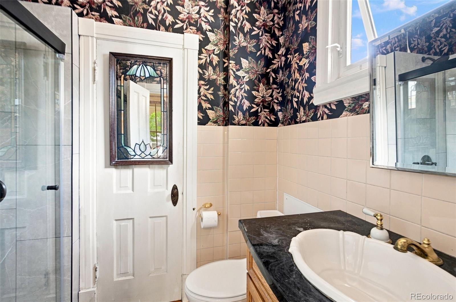 Bathroom with floral wallpaper, a stained-glass door panel, and a black stone vanity.