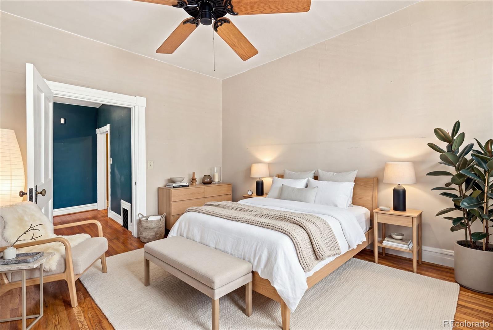 Staged bedroom with a white bed, hardwood floors, and an open doorway to a blue-painted room.