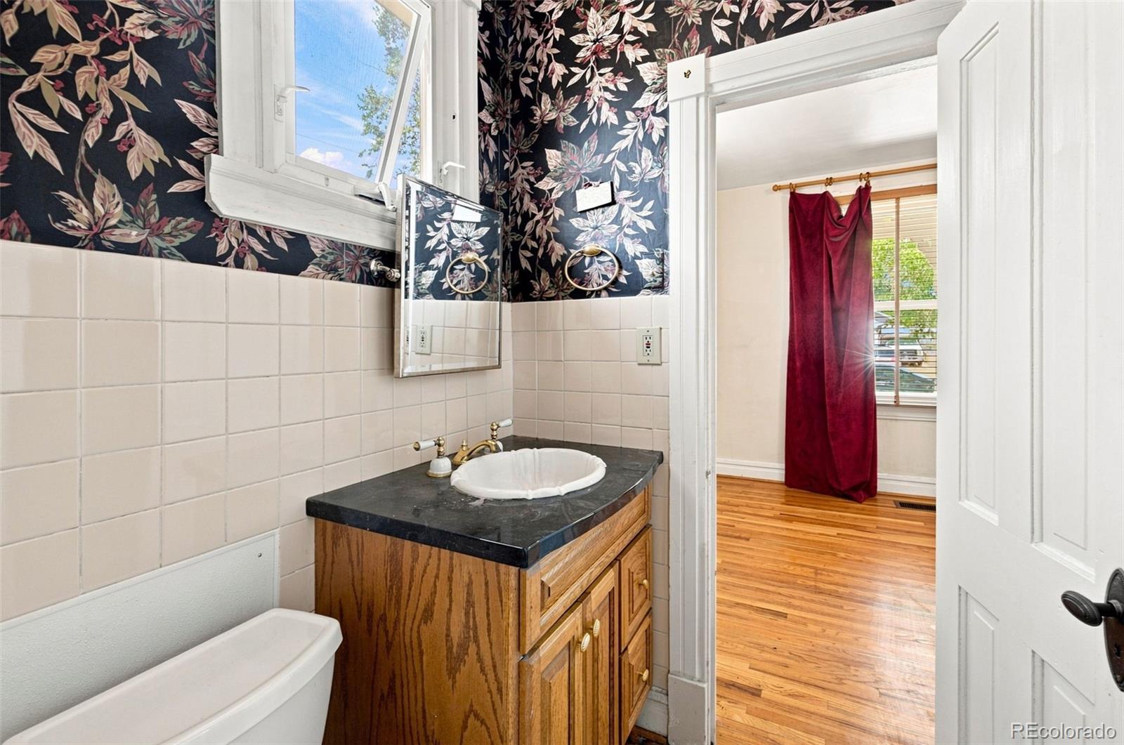 Bathroom with floral wallpaper, a wood vanity, and an open doorway to a hardwood-floored room.