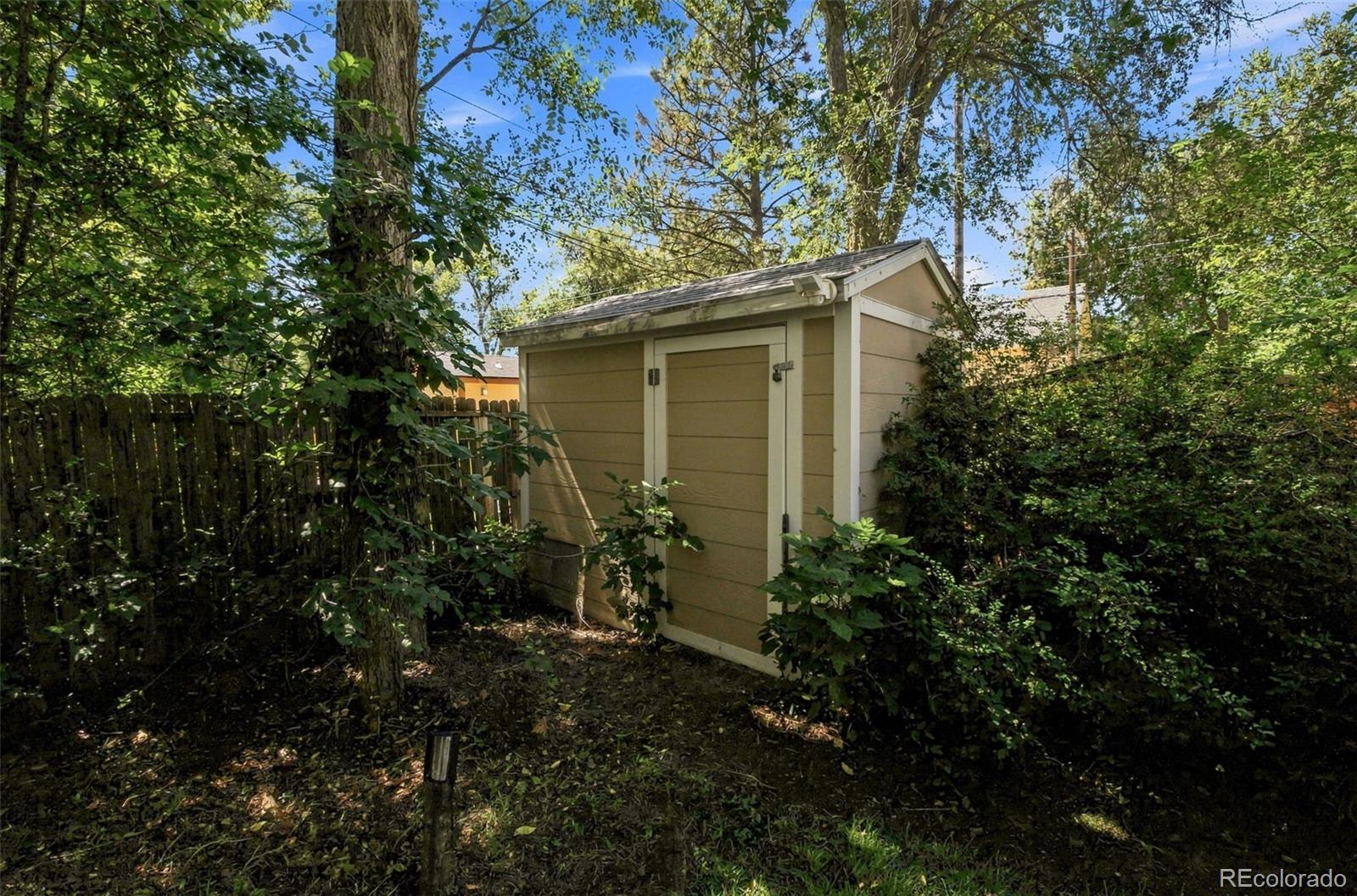 Tan storage shed with white trim nestled among mature trees and shrubs in a shaded backyard.