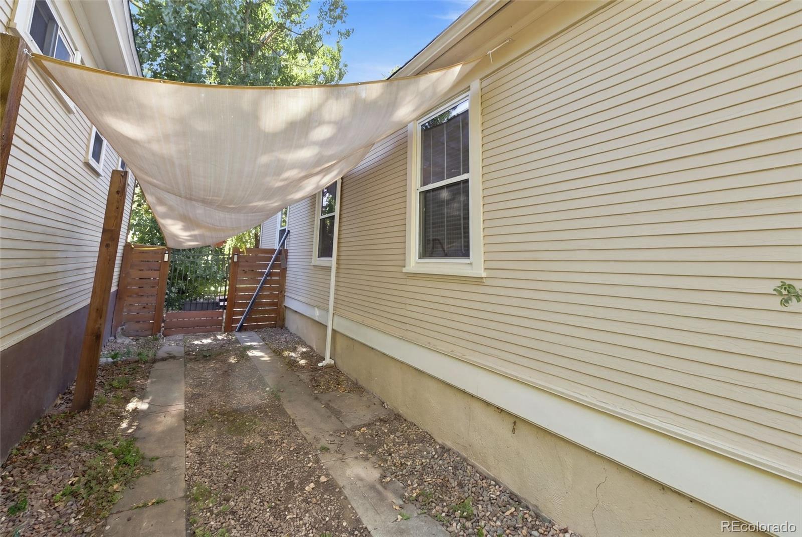 Side patio with a fabric shade sail, gravel walkway, and wooden gate.