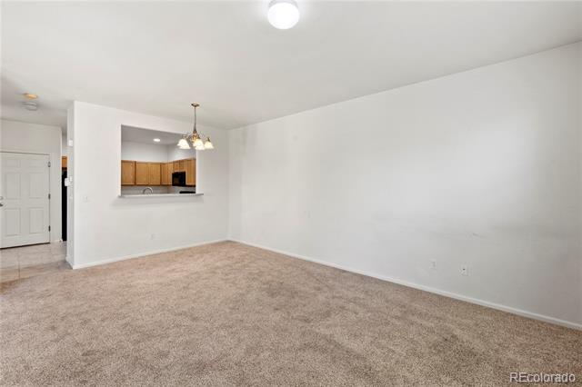 Empty carpeted living room with a wide kitchen pass-through and the entry door at left.