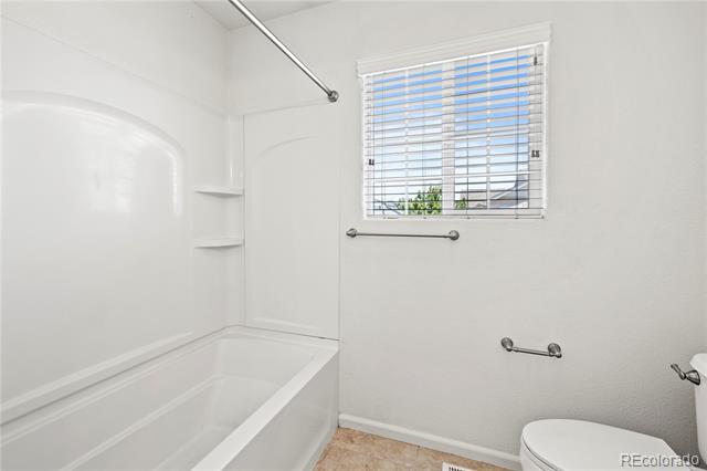 White bathroom with a bathtub, toilet, grab bars, and a window with blinds.