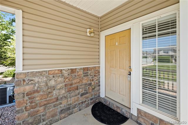 Covered home entry with tan siding, stone veneer, a paneled door, and a large window with blinds.
