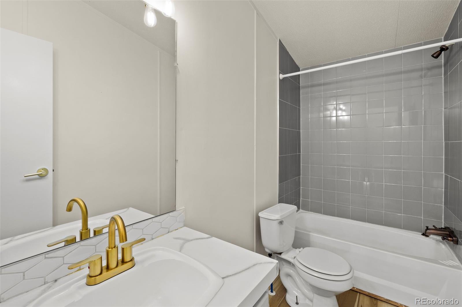 Bathroom with a white vanity, gold-tone faucet, toilet, and bathtub against gray tile.