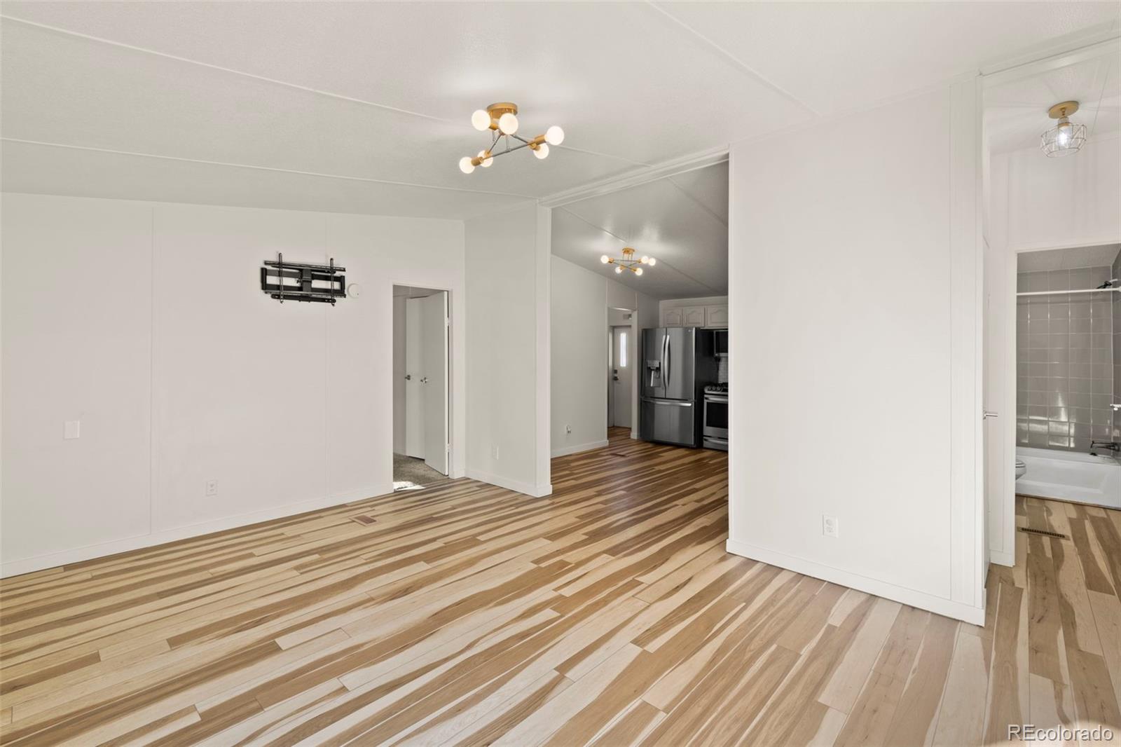Empty living area with striped wood flooring opening to a kitchen and bathroom.