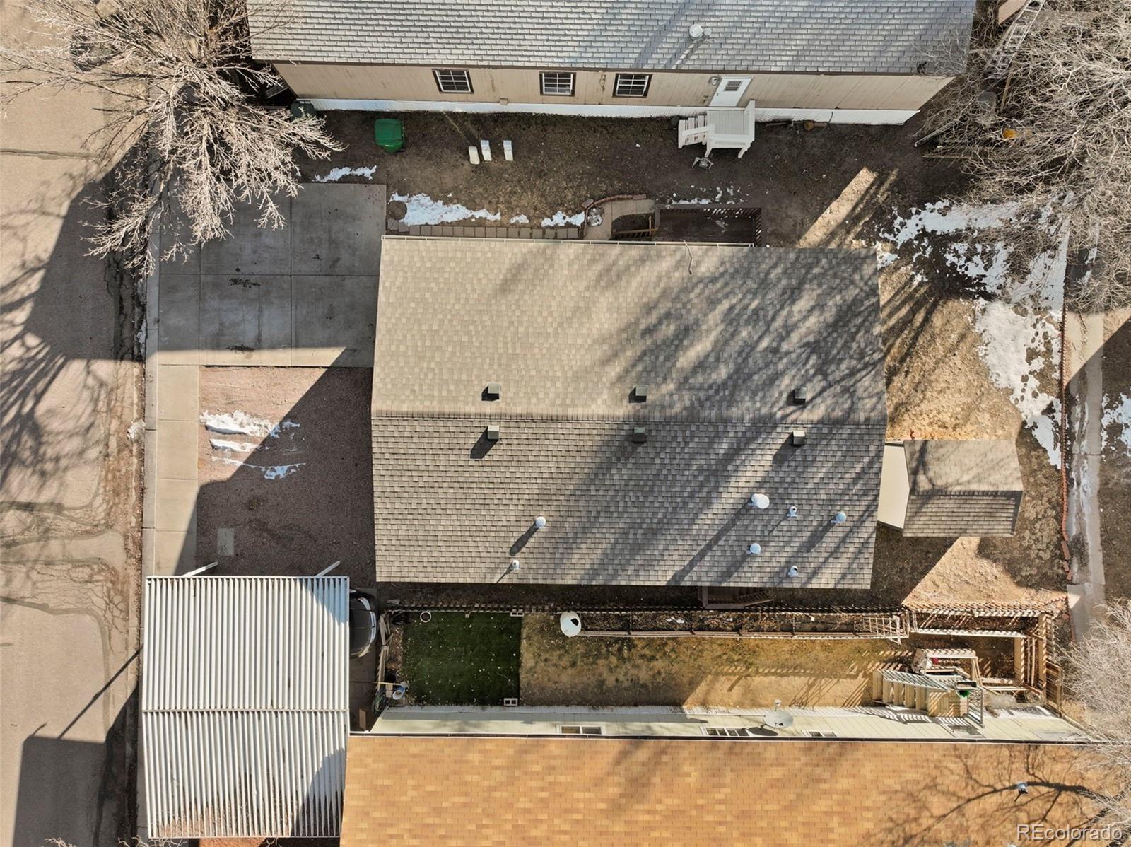 Aerial view of a home with a large shingled roof, driveway, fenced yard, and scattered snow.