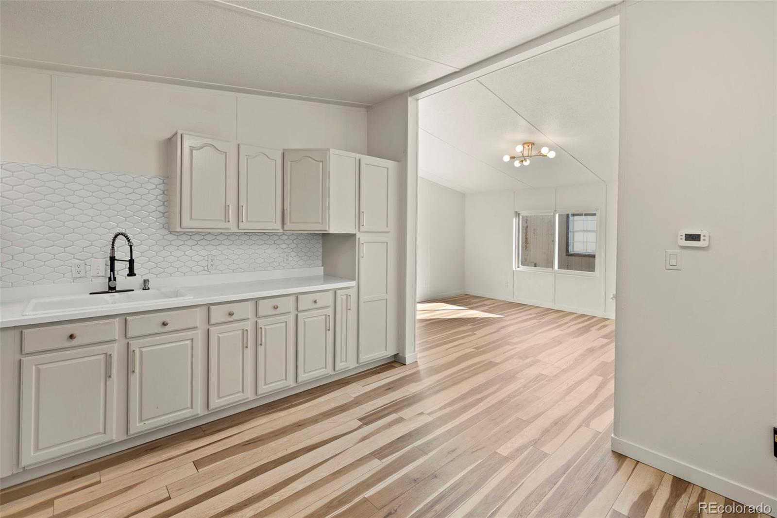 Vacant kitchen with white cabinets, hexagonal tile backsplash, and light plank flooring opening to a sunny living area.