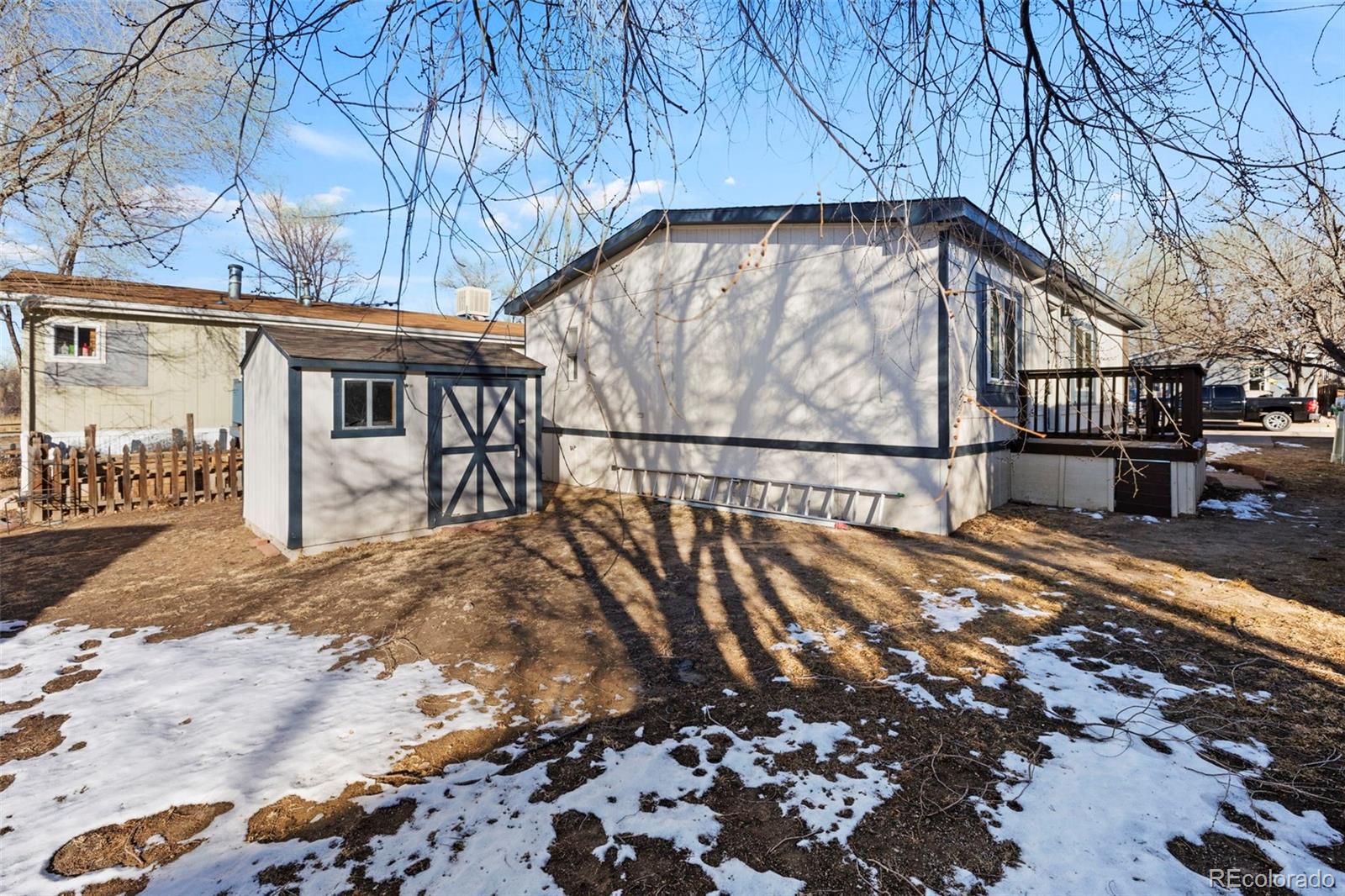 Rear of a light-colored house with a raised deck and detached shed in a snow-patched yard.