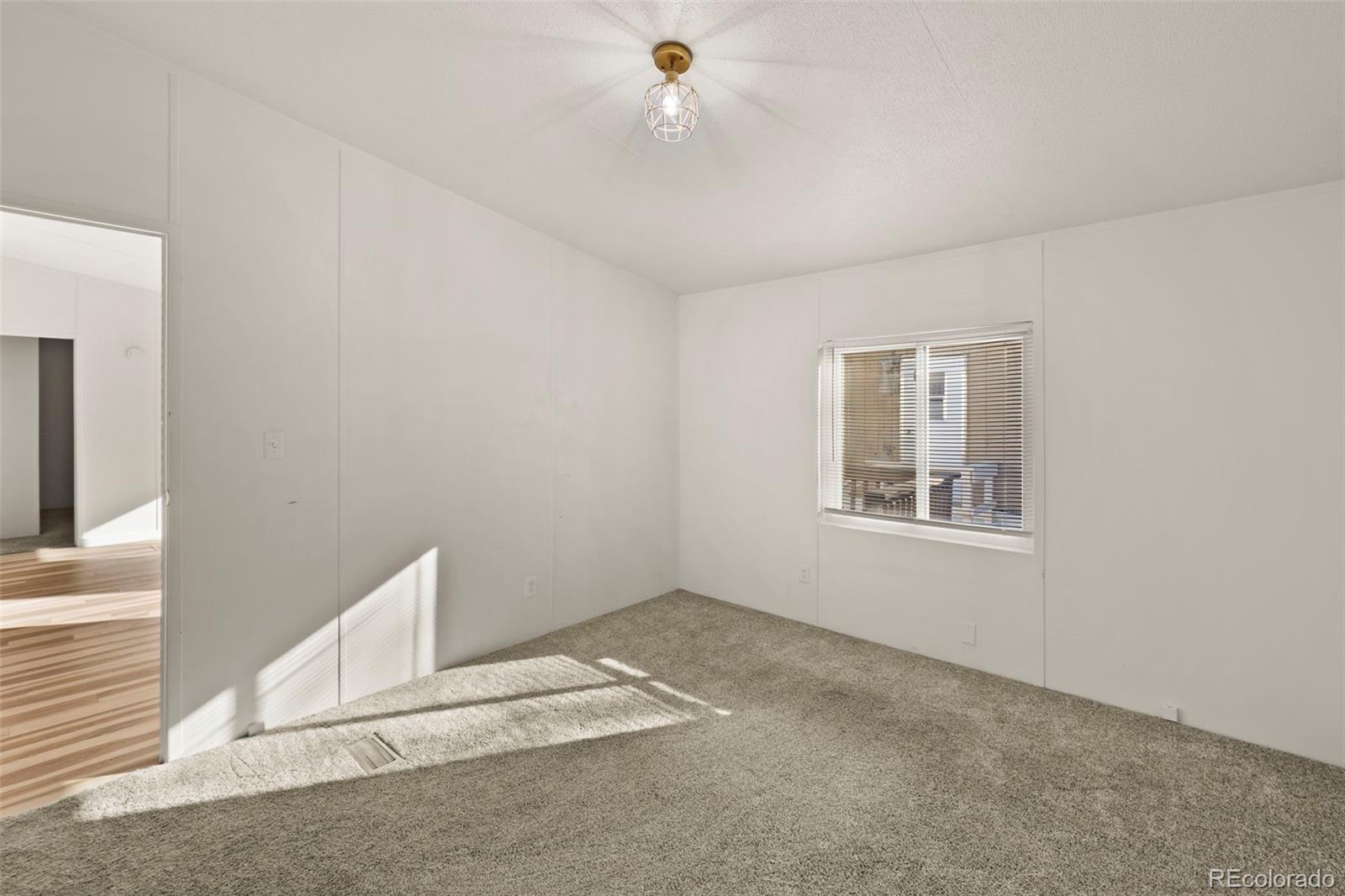 Empty carpeted bedroom with white walls, a window with blinds, and sunlight across the floor.