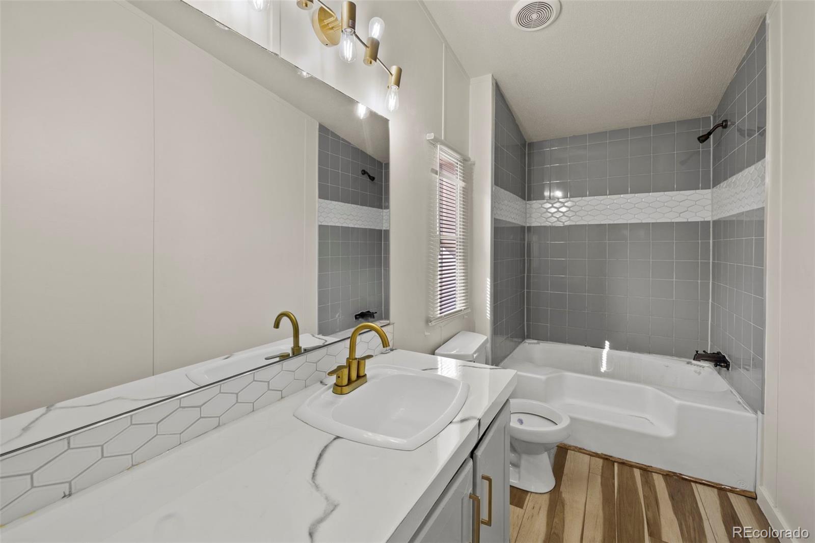 Bathroom with a white vanity, gold faucet, gray tiled walls, and bathtub.