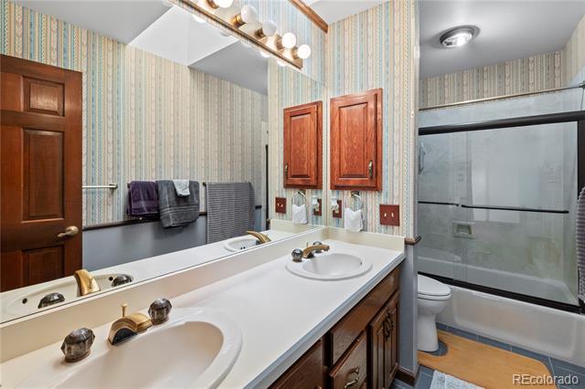 Bathroom with a double-sink vanity, wood cabinets, striped wallpaper, and a tub-shower enclosure.
