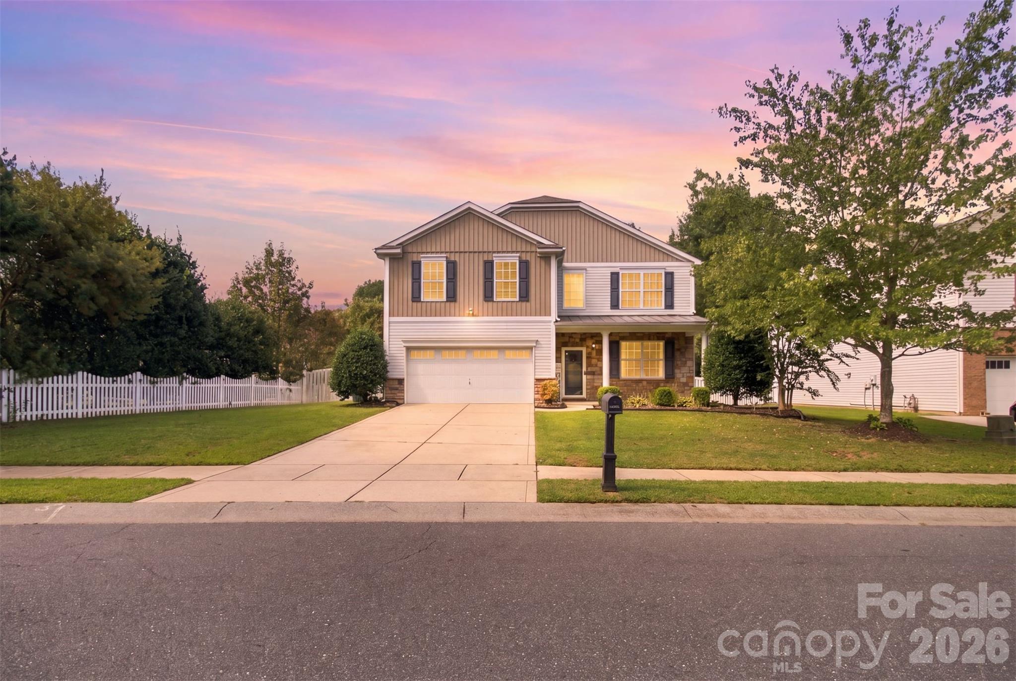 Two-story house with a wide driveway, front lawn, white fence, and glowing windows at dusk.