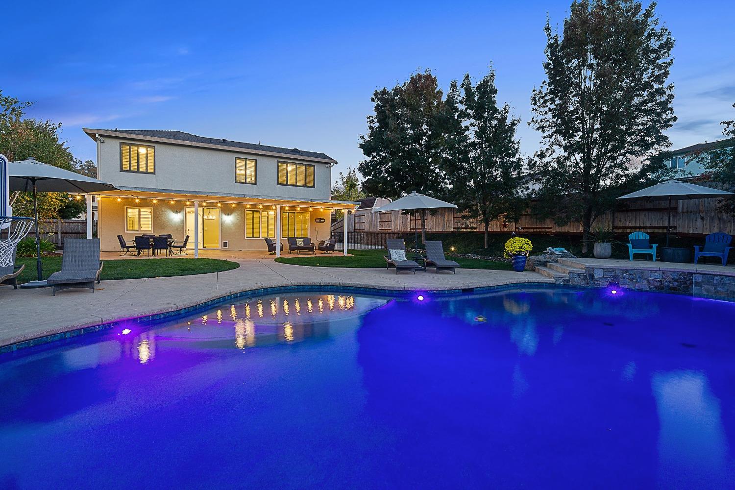 Backyard pool and two-story home at dusk, with illuminated water and a covered patio strung with lights.