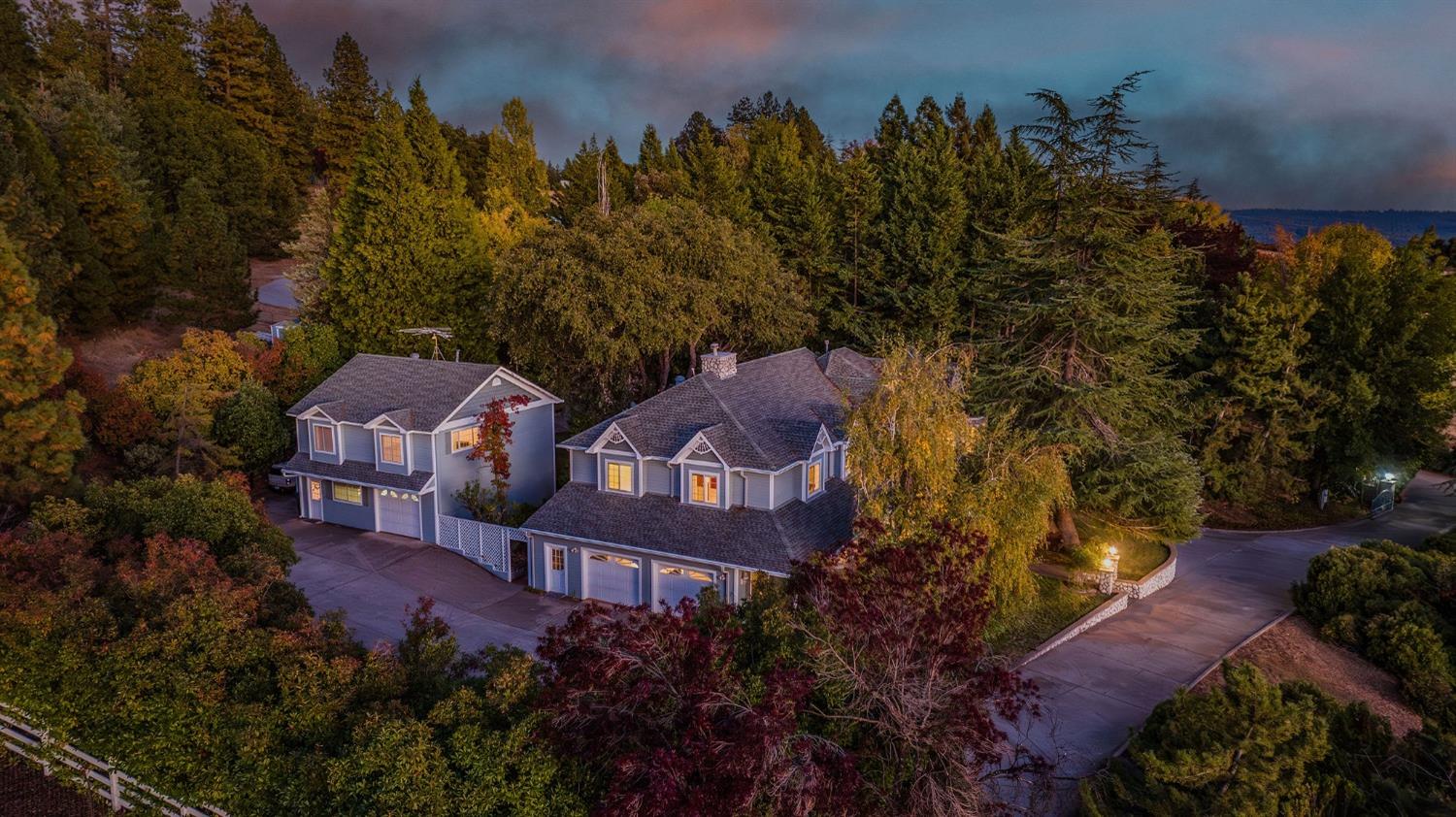 Aerial view of two homes among autumn trees at dusk, with exterior lights glowing.