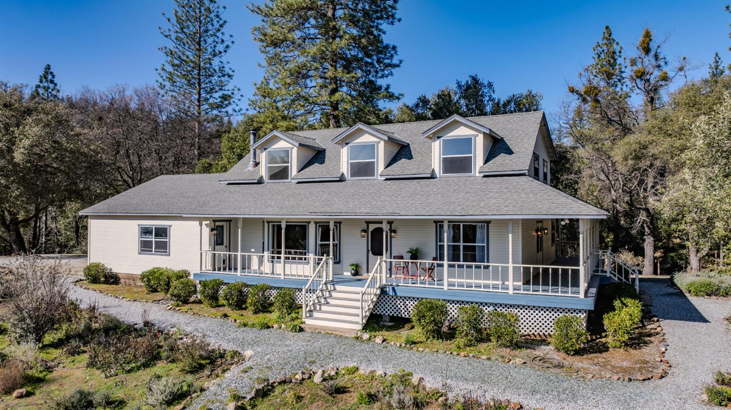 Light-colored home with three dormers, a wraparound porch, and a gravel driveway among trees.