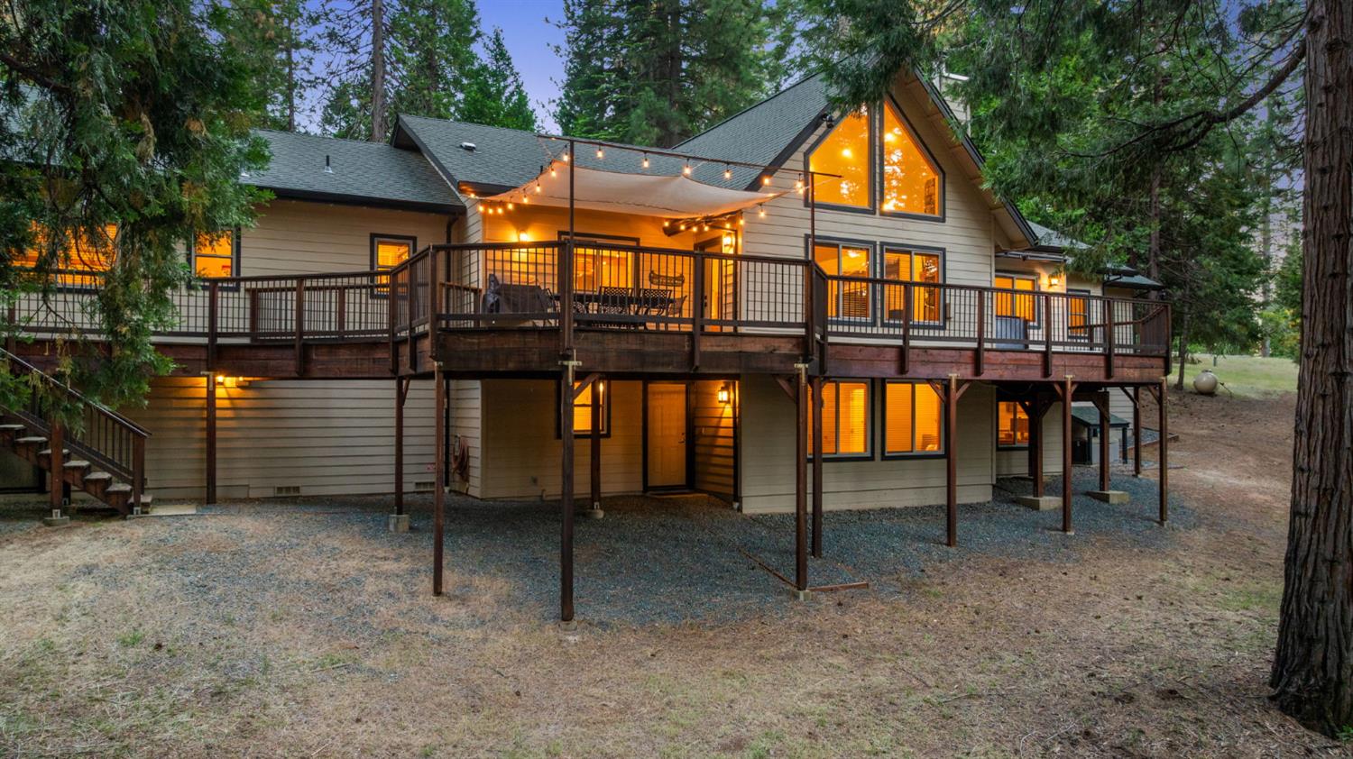 Two-story home with a large elevated deck, shade sail, and glowing lights among tall trees at dusk.
